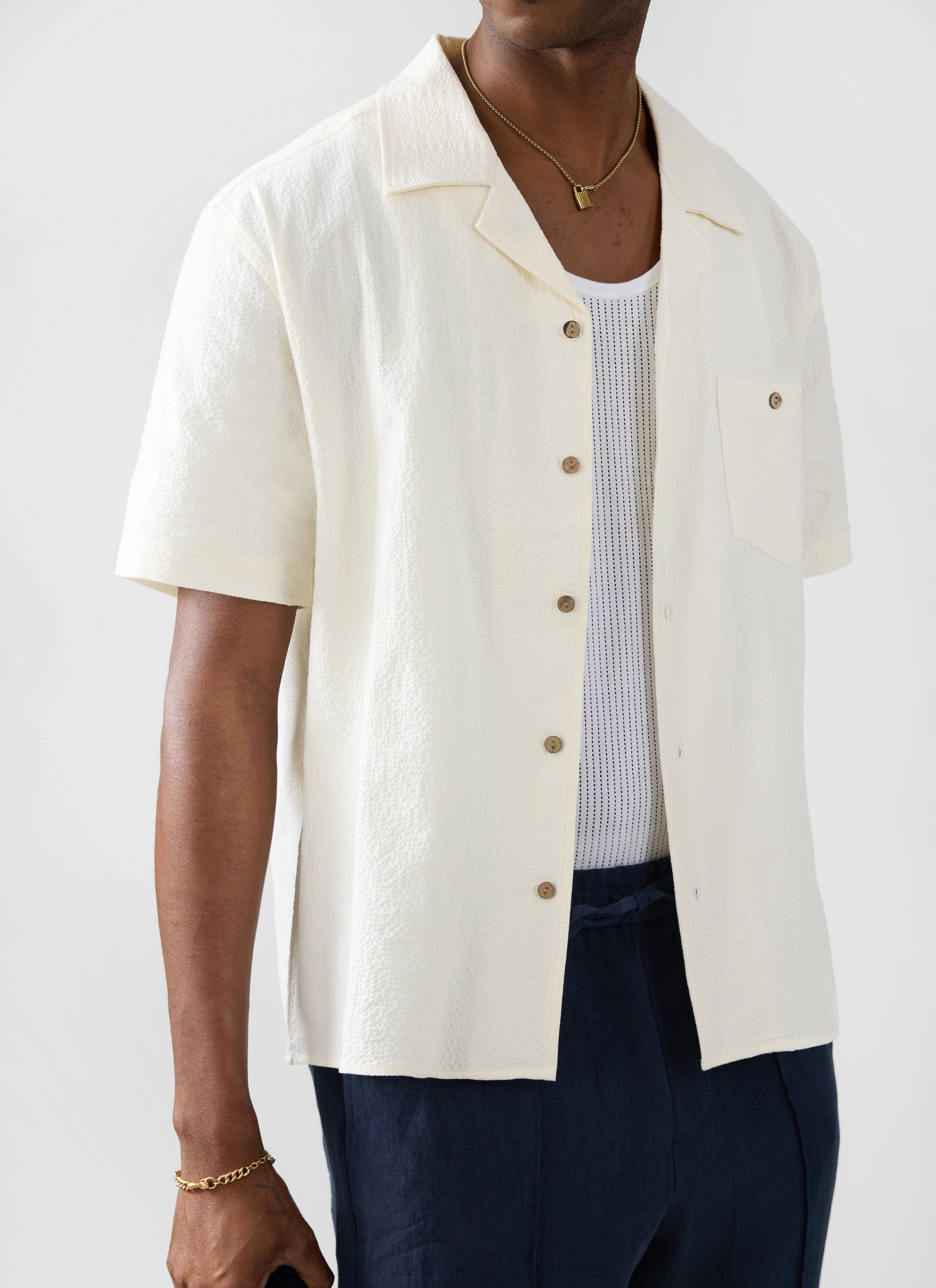 Model wears the Percival ecru seersucker Cuban Shirt open over a white vest and styled with navy trousers and gold jewellery.