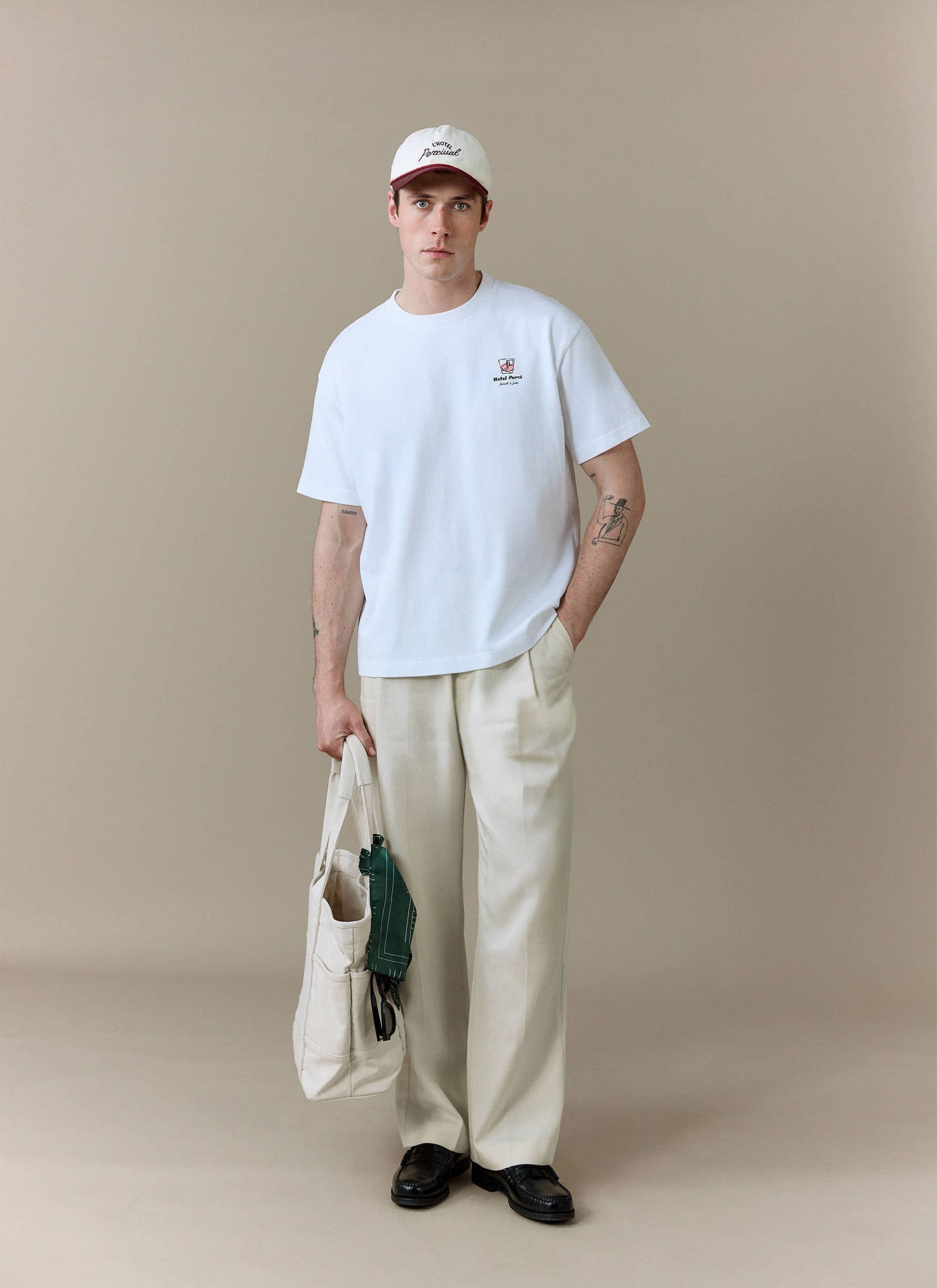 A model wears the white Hotel Bistro Oversized T Shirt from Percival, styled with cream trousers, a cap and a tote bag.