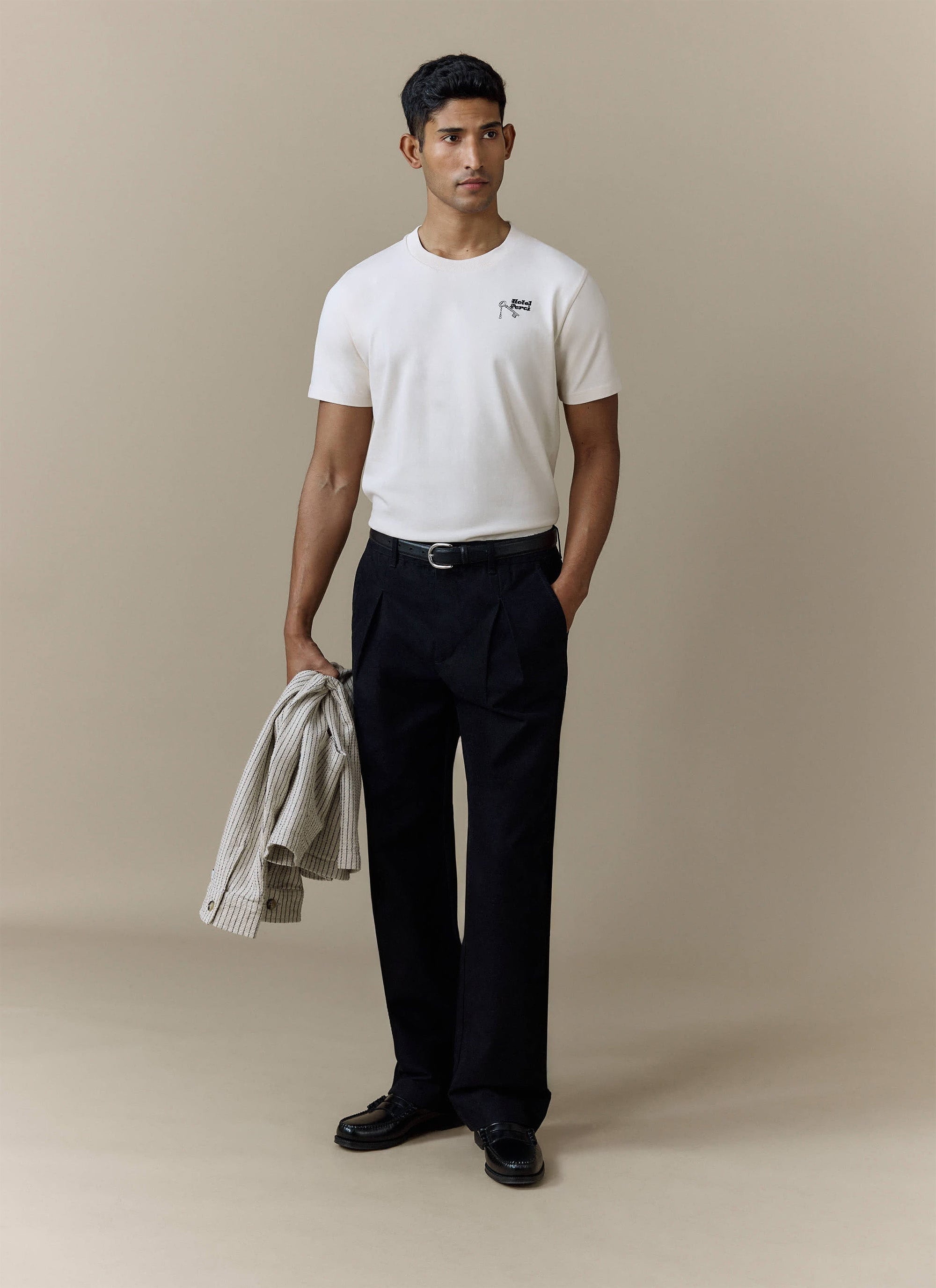 A man models the Percival Hotel T Shirt in ecru, holding a striped jacket and wearing smart black trousers.