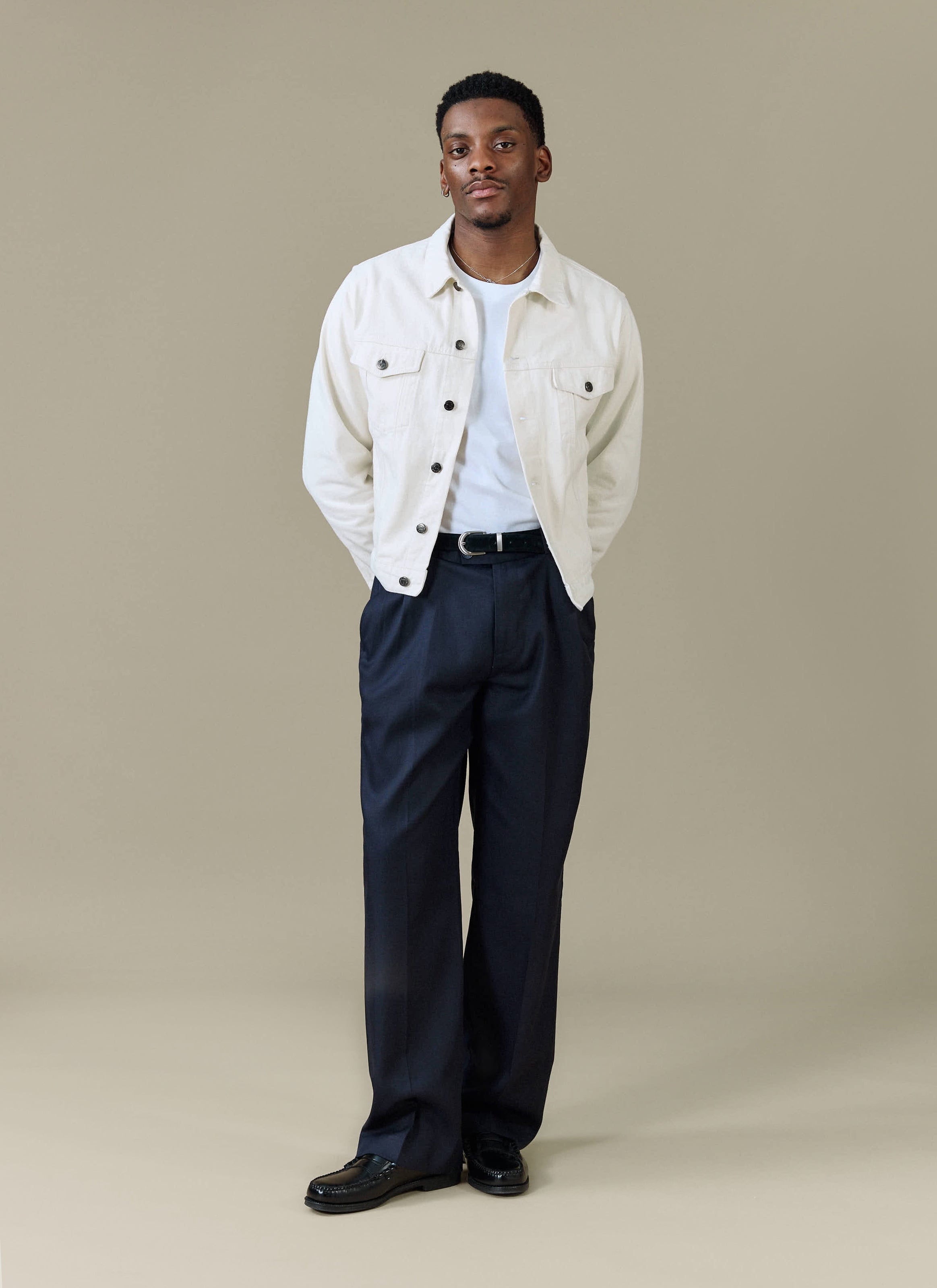 A man models the navy Pleated Leg Trousers from Percival with a white t-shirt and cream jacket.