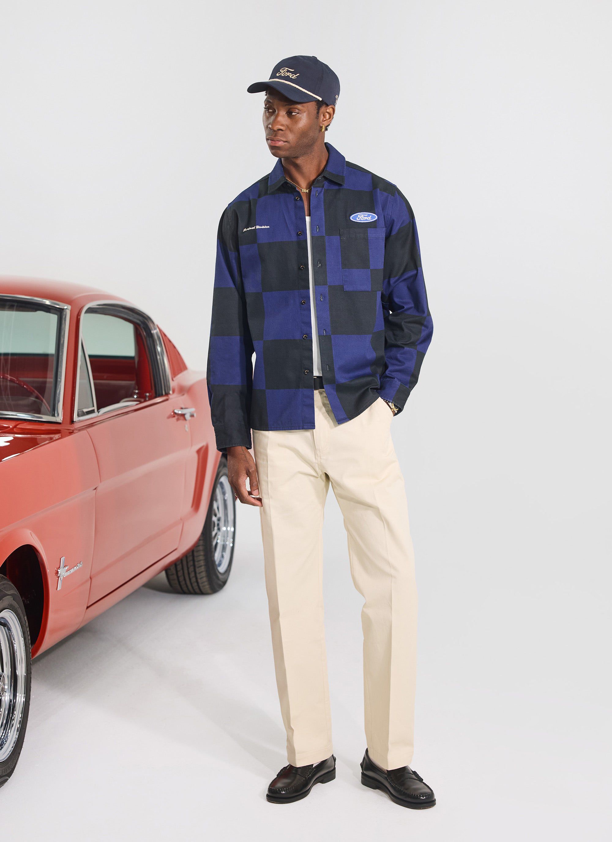 A model wears the Percival x Ford navy and black checkerboard printed overshirt, standing next to a classic red Mustang.