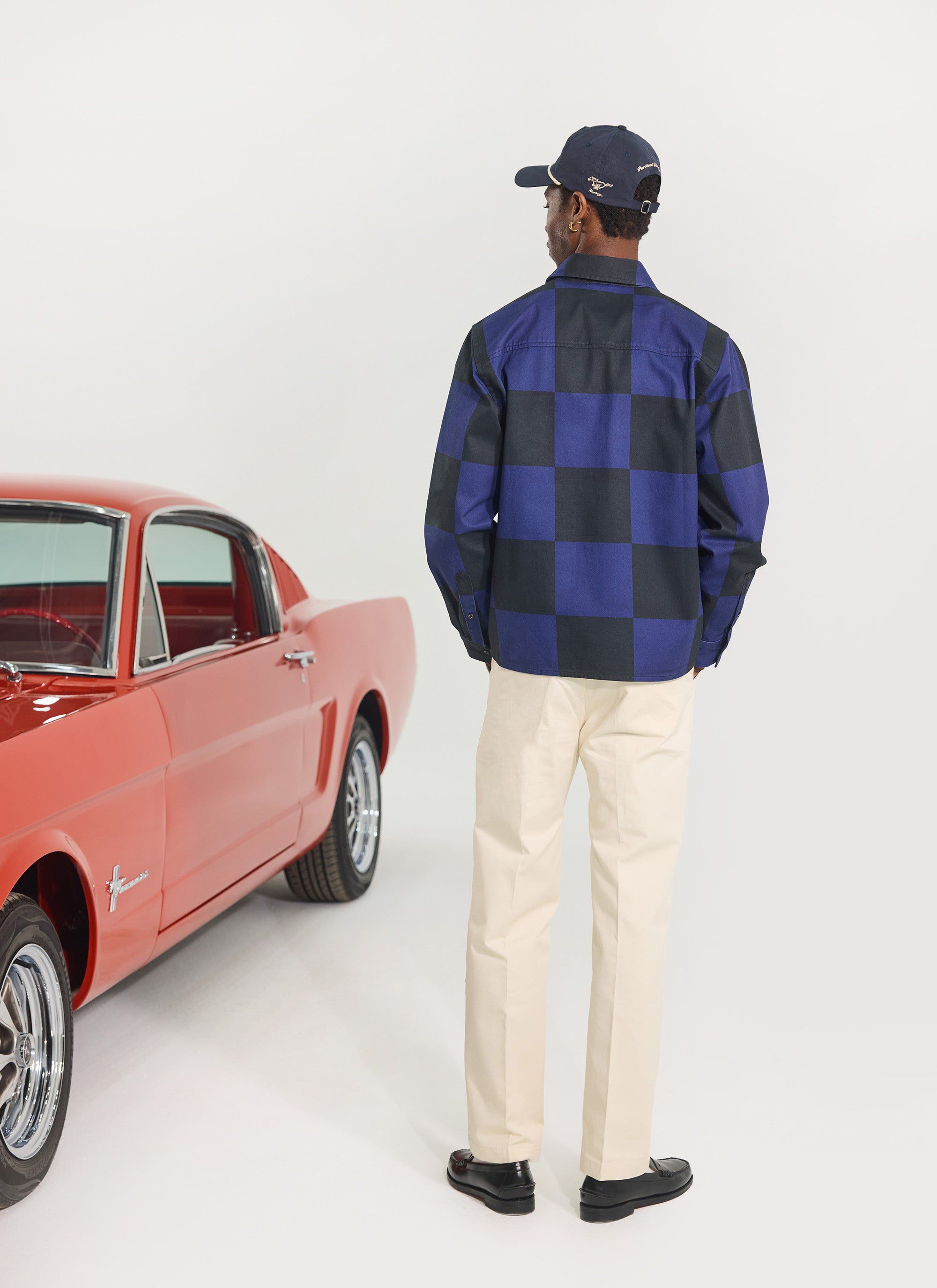 A model from behind wears the Percival x Ford navy and black checkerboard overshirt next to a classic red Ford Mustang.
