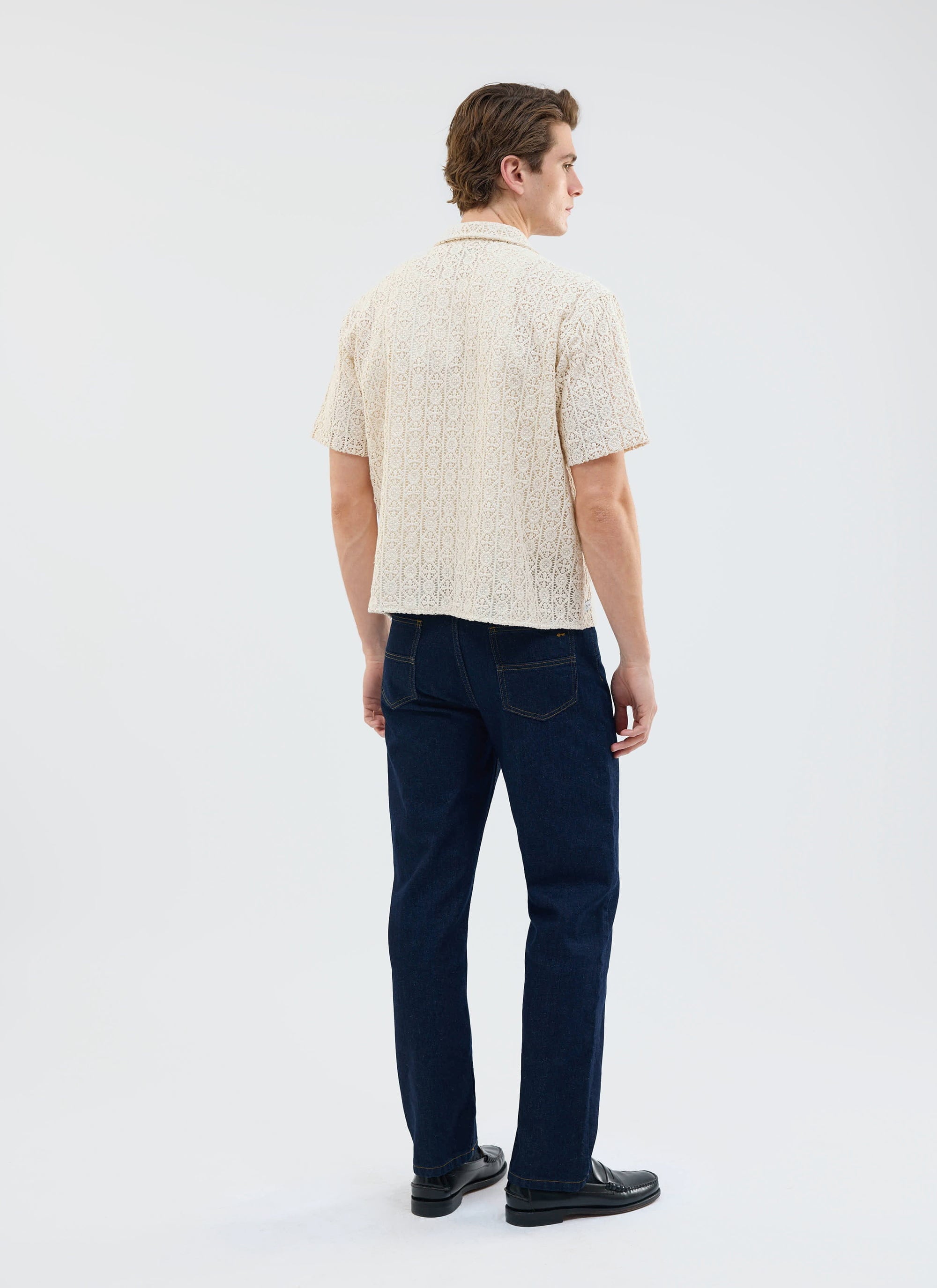 A model, seen from behind, wears the Percival Elio Crochet Cuban Shirt in ecru with dark wash jeans and black loafers.