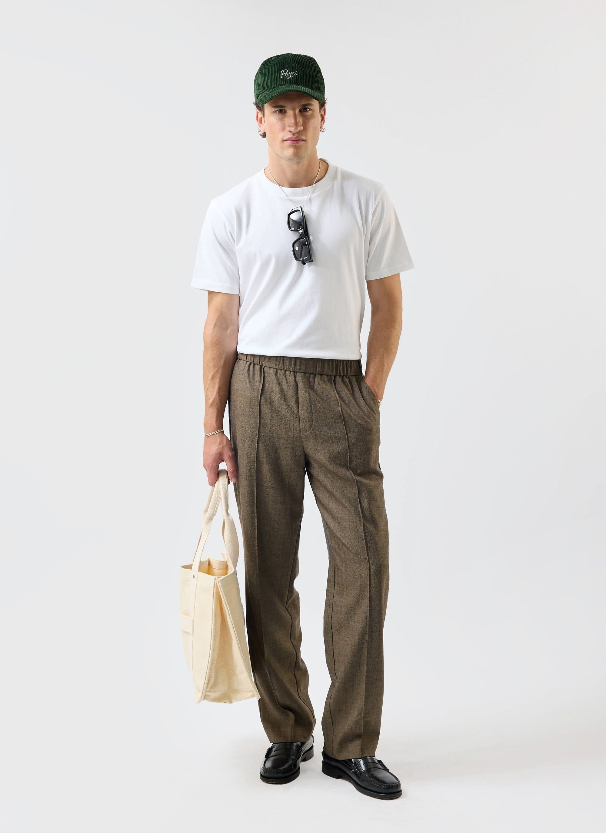 A man models the relaxed fit Percival Grounds Trousers in oak herringbone wool, paired with a white tee and loafers.