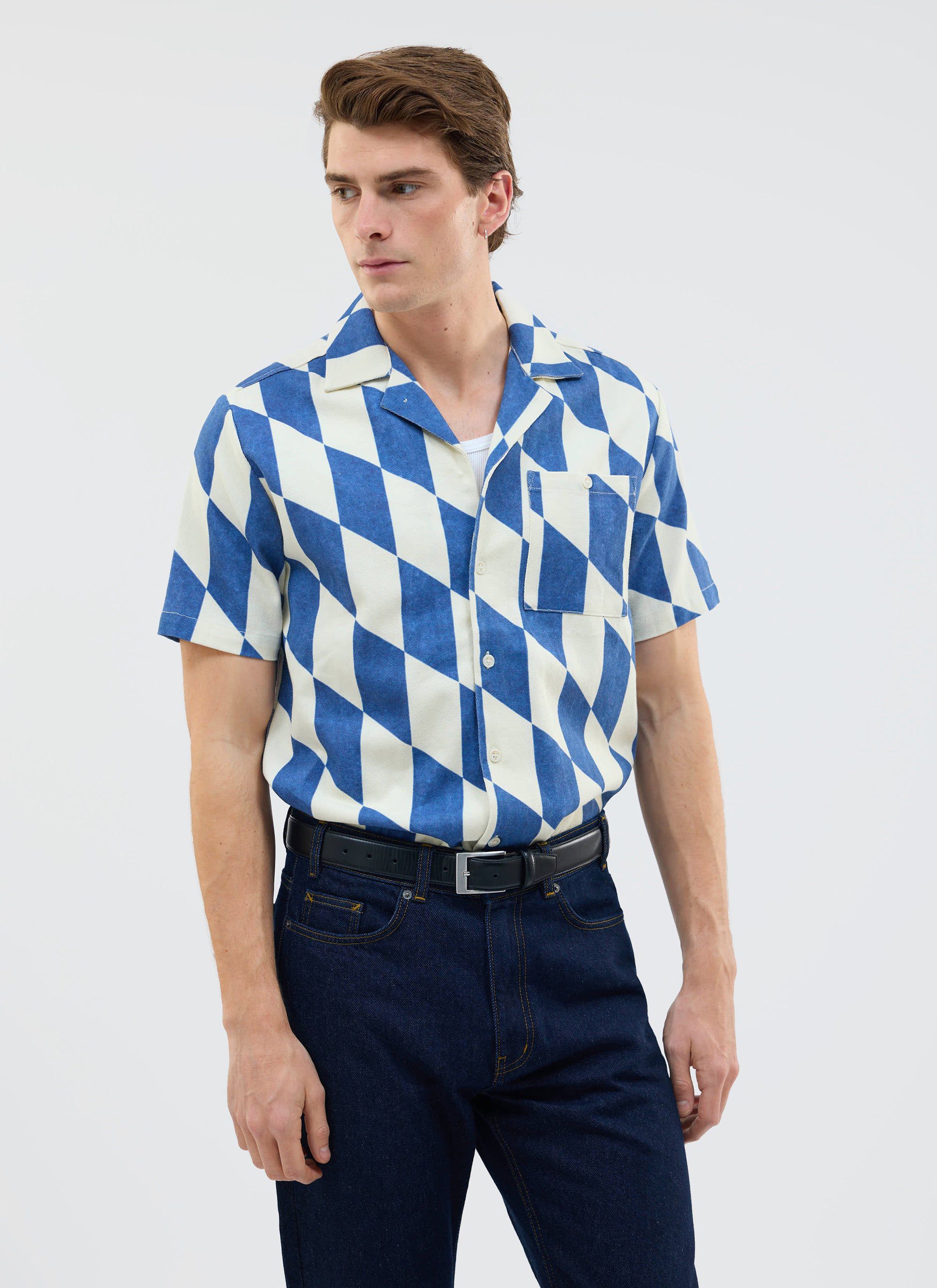 A model wears the Percival Market Diamond Cuban Shirt in ecru and navy, which has a diamond pattern, with dark denim jeans.