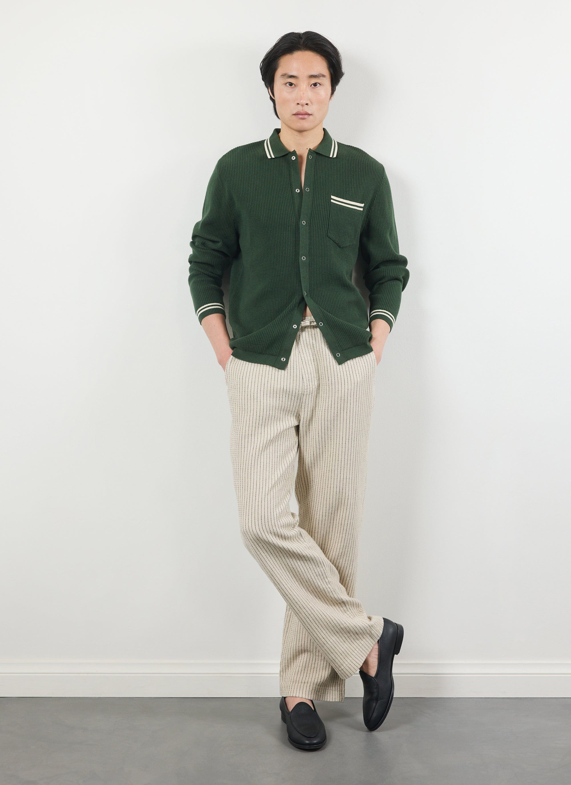 A model styles the ecru Percival Sorrento Linen Trousers with a green knit cardigan and black loafers against a white wall.