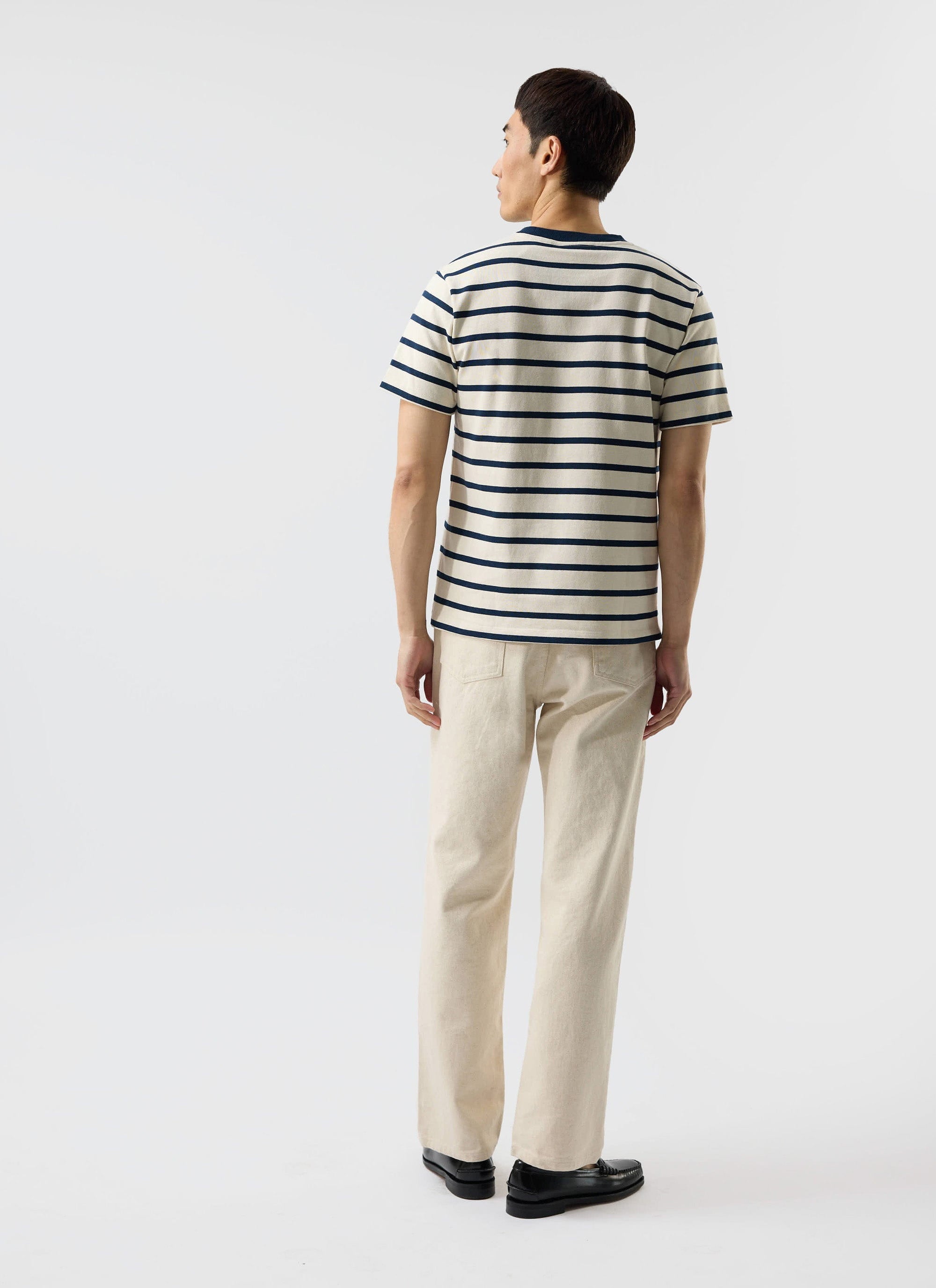 A model seen from behind wearing the Percival Breton Stripe T-shirt in ecru and navy with cream trousers and black loafers.