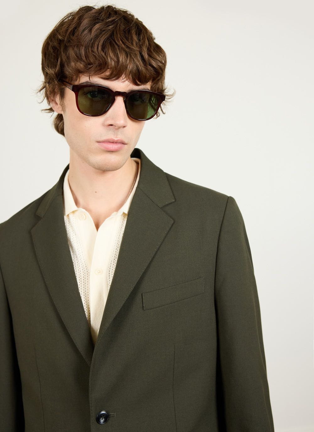 A model wears the Percival Tailored Blazer in Forest Green wool, styled with a cream polo shirt and sunglasses.