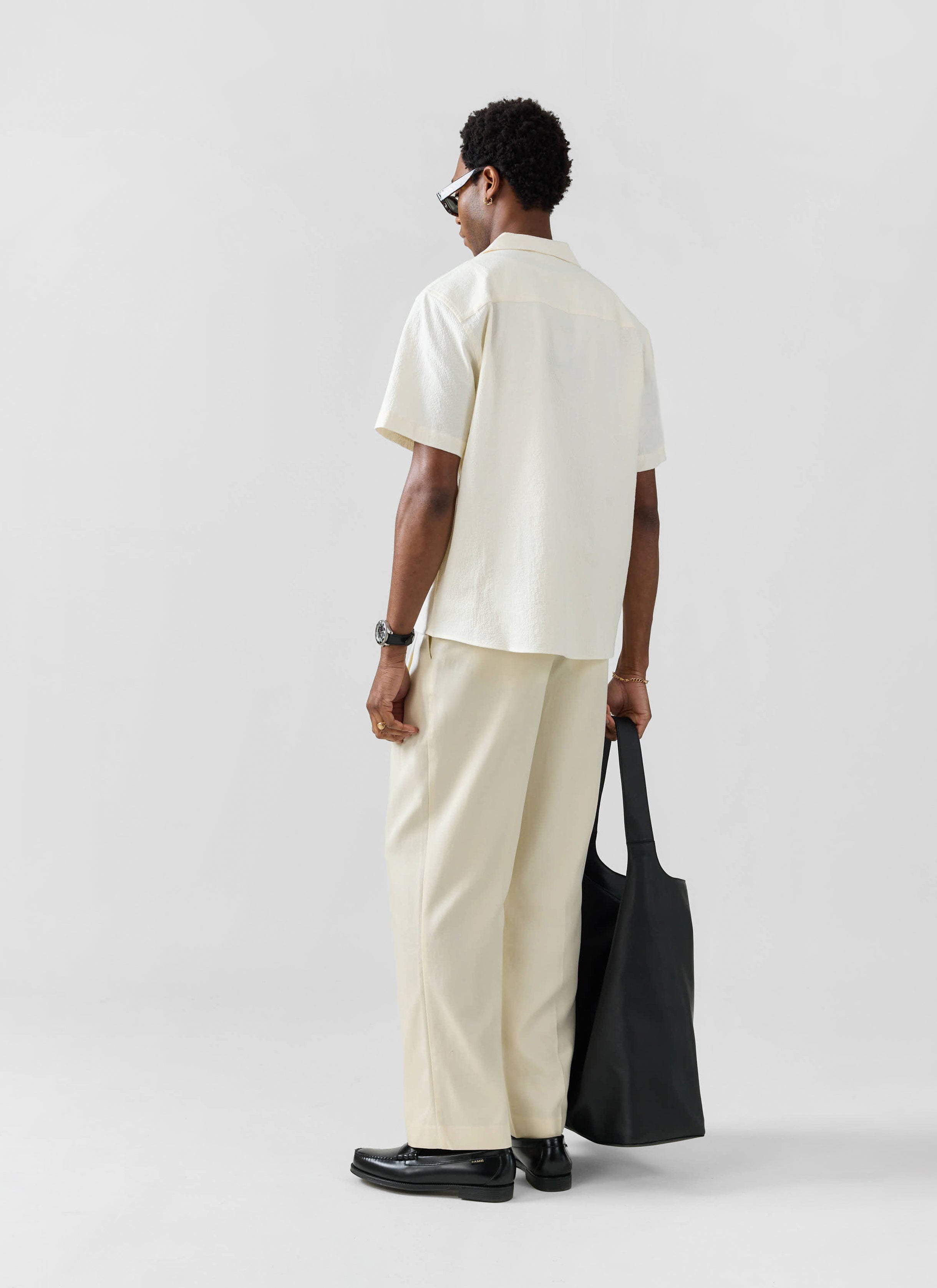 Back view of a model wearing the Percival ecru seersucker Cuban shirt and matching trousers with a black tote bag.