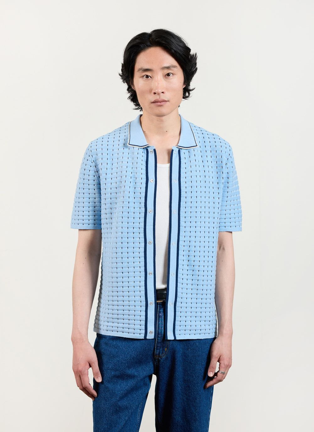 A model wears the Percival Broken Stripe Knitted Shirt in blue, styled open over a white vest and blue jeans.