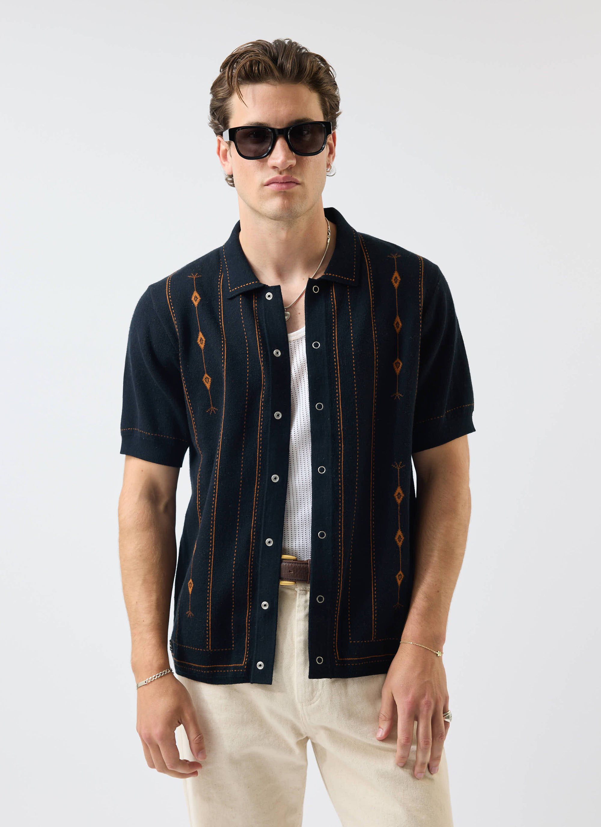 A model wears the Percival Folio Knitted Shirt in dark navy, styled open over a white vest with cream trousers.