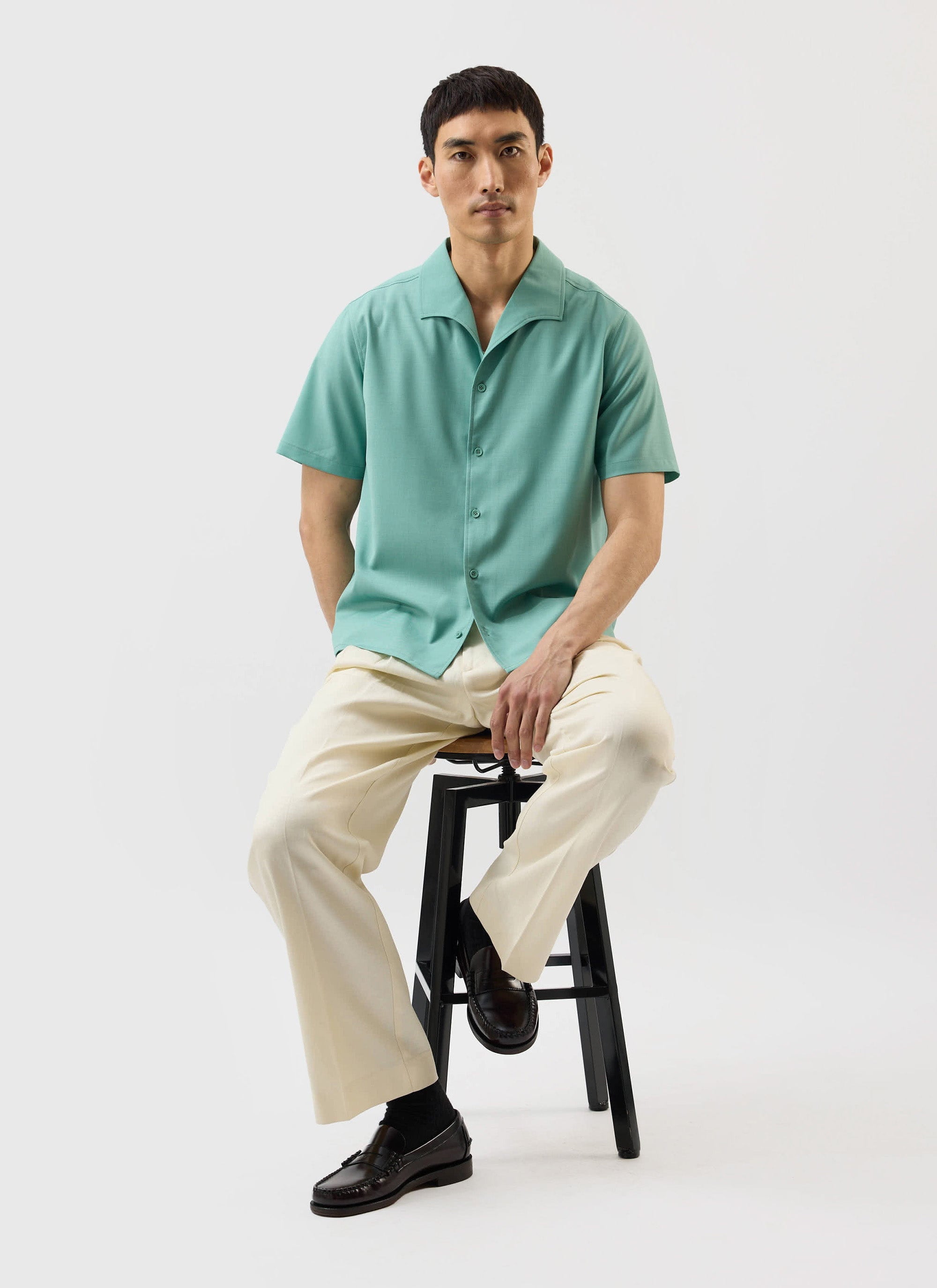 A model sits on a stool wearing the Percival x Warren Drape Cuban Shirt in mint green with cream trousers.