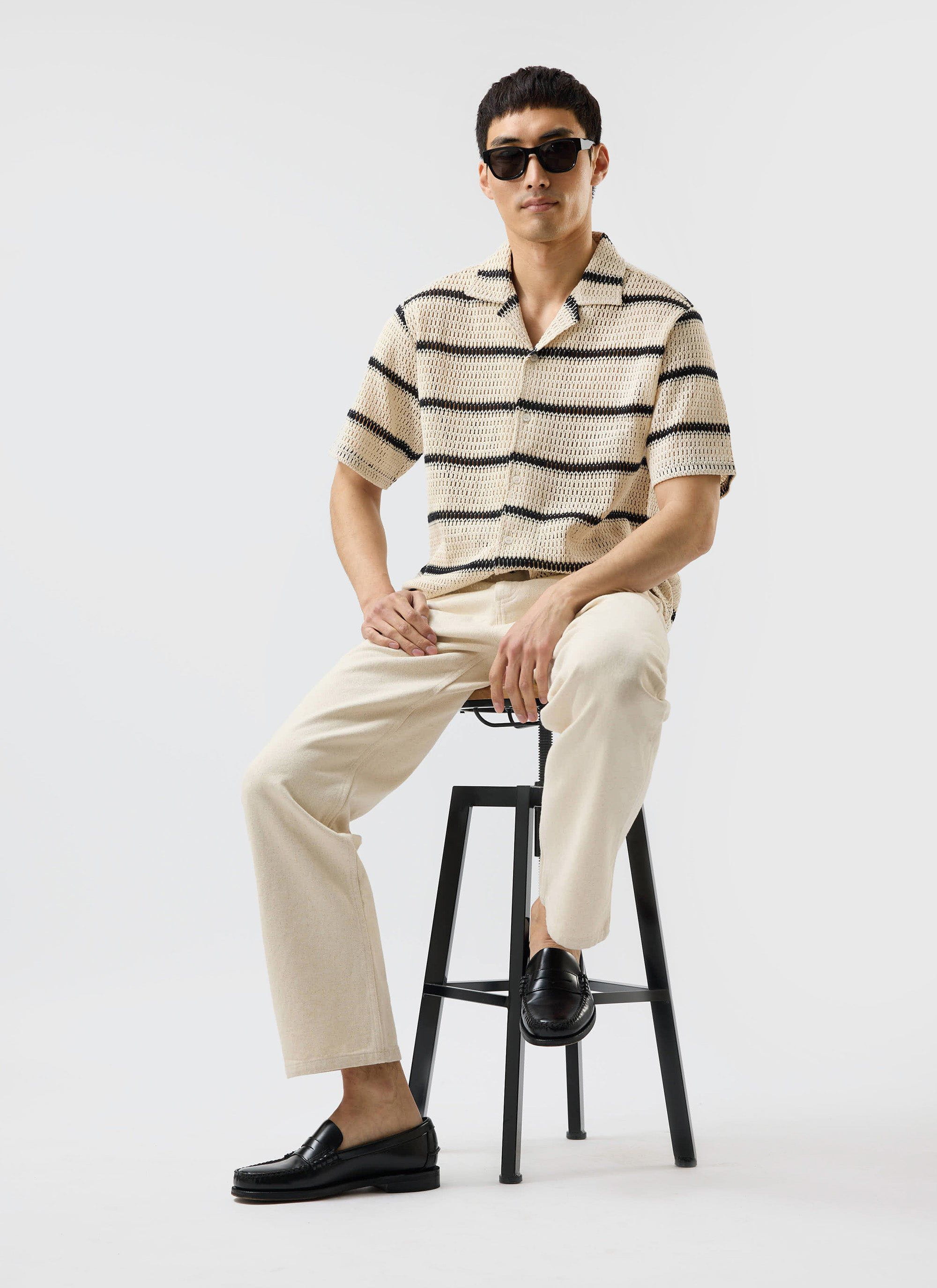 A model sits on a black stool, wearing the Percival Crochet Groundsman Cuban Shirt in ecru with black stripes.