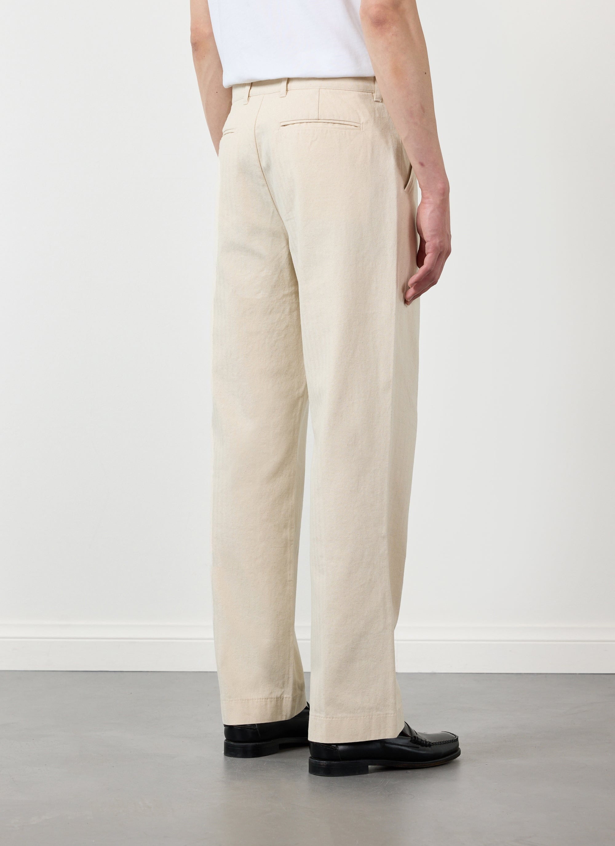 Back view of a model wearing Percival's ecru cotton Program Pleated Trousers with a white t-shirt and black loafers.