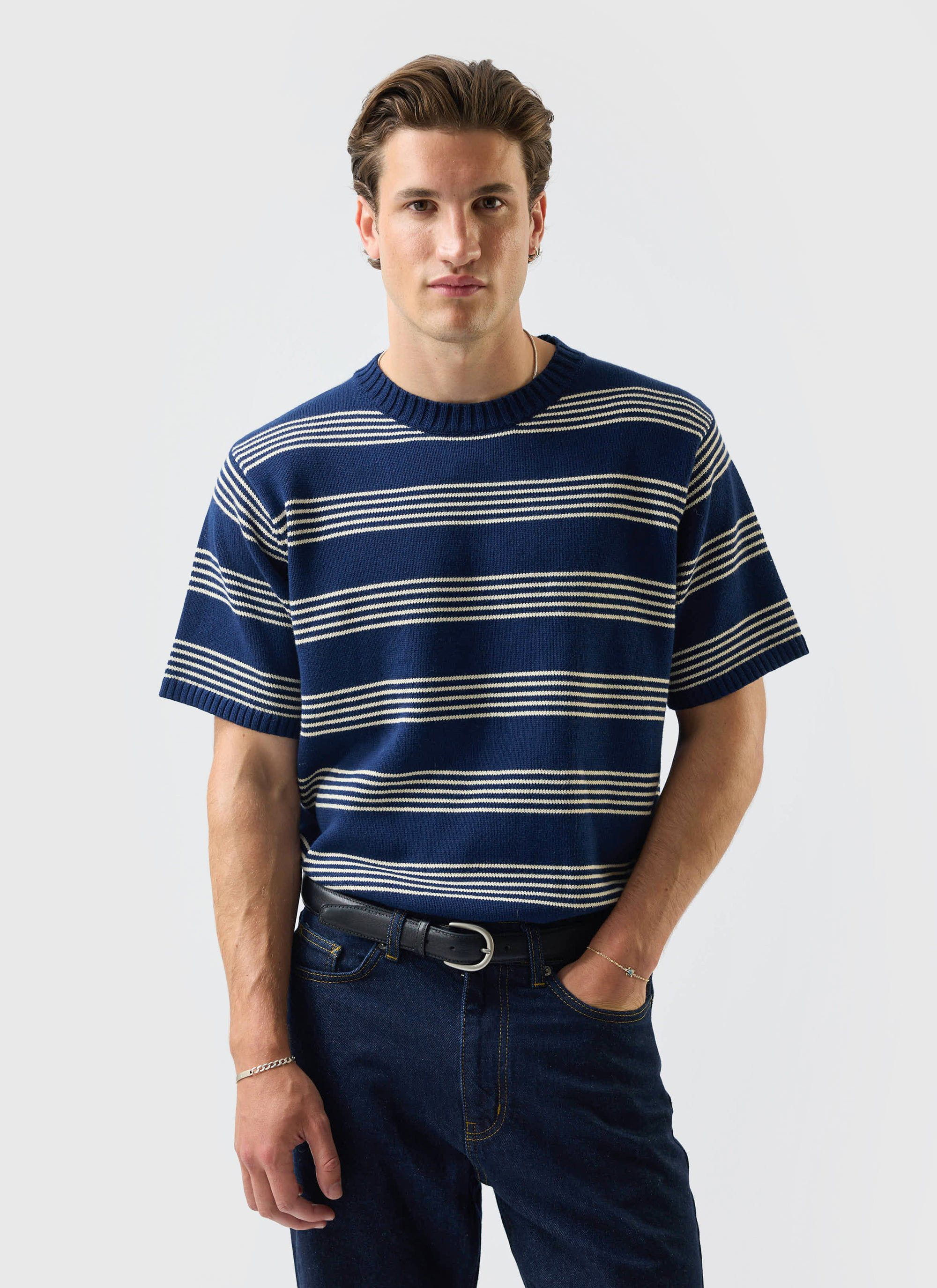 A model wears the Percival Fiction knitted t-shirt in navy with cream stripes, paired with dark denim against a white wall.