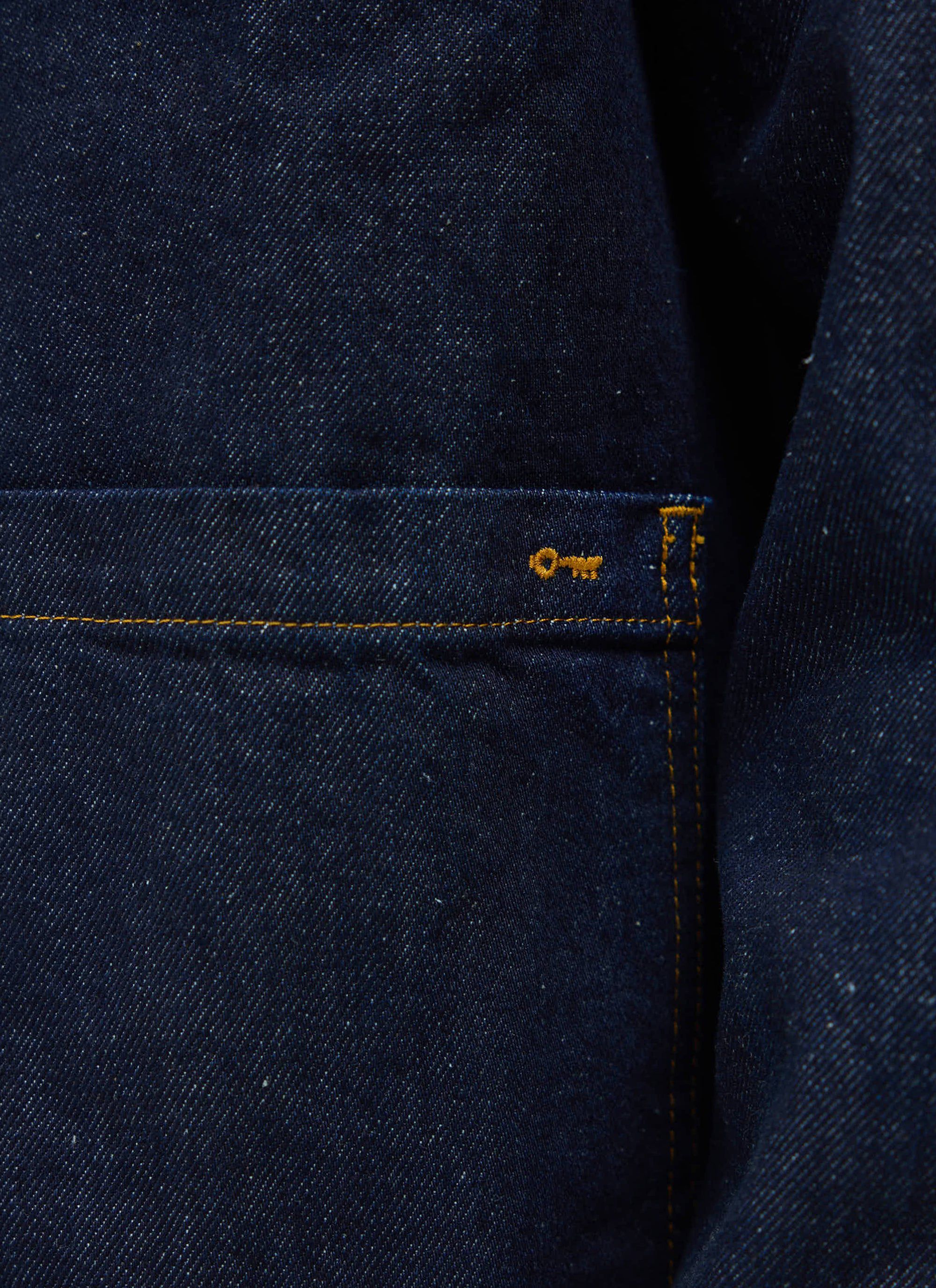 A close-up of the Percival Gove Chore Jacket in indigo denim, showing the patch pocket with contrast stitching and key logo.