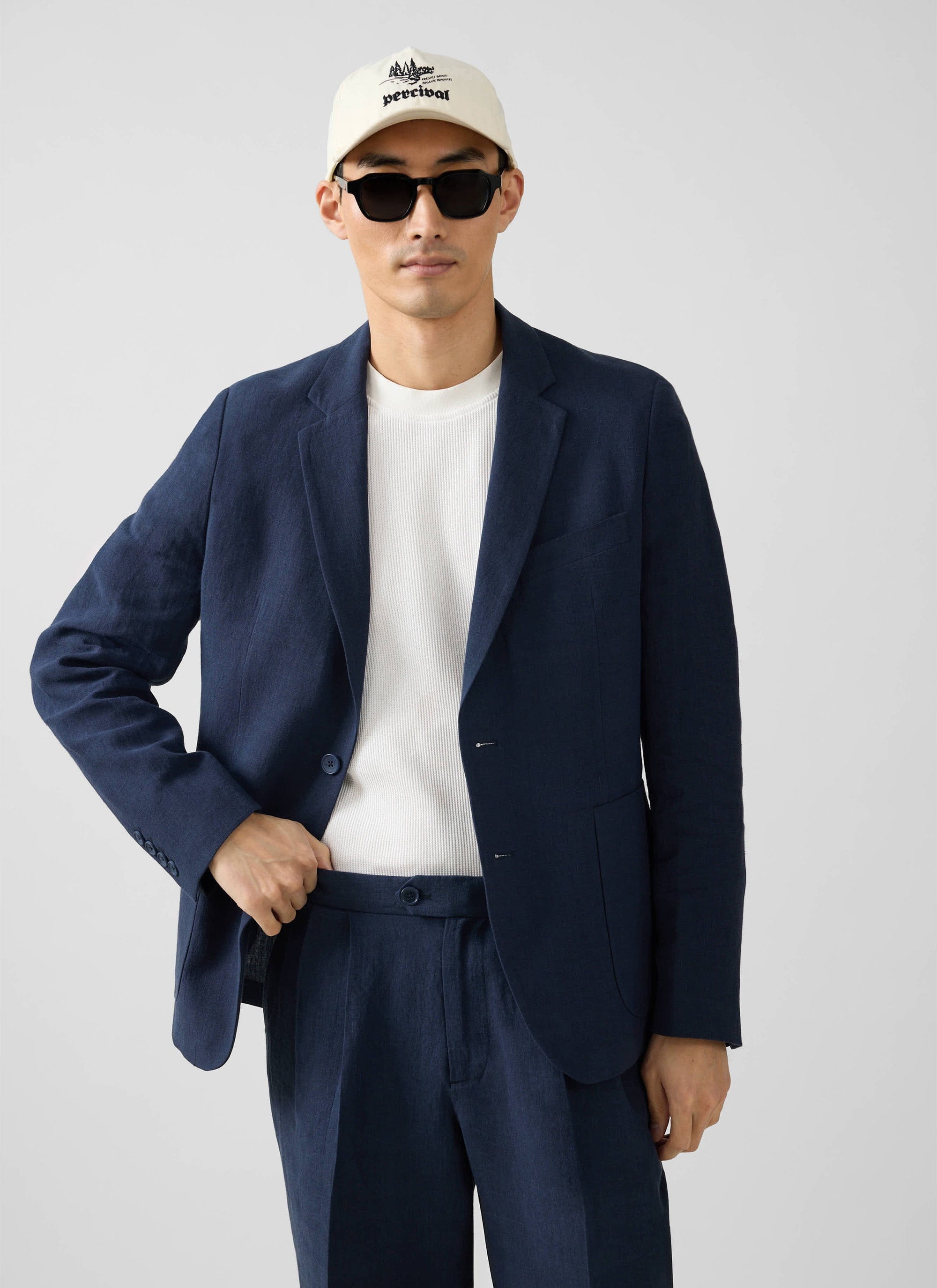 A man models the Percival navy linen suit, styled with a white t-shirt, sunglasses and a Percival cap