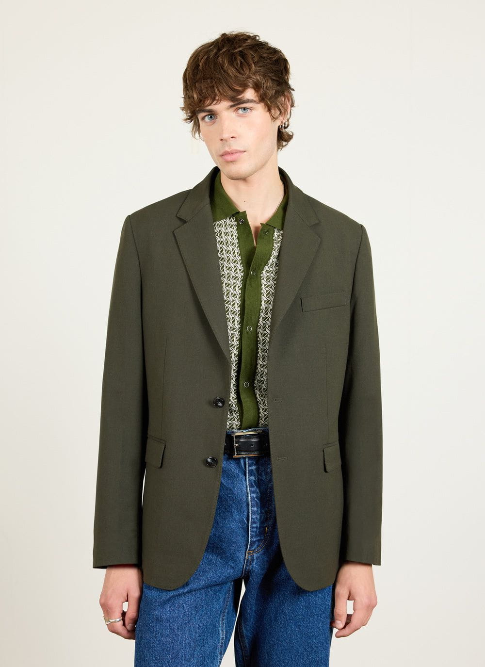 A man models the Percival forest green wool tailored blazer with a patterned green polo and blue jeans.