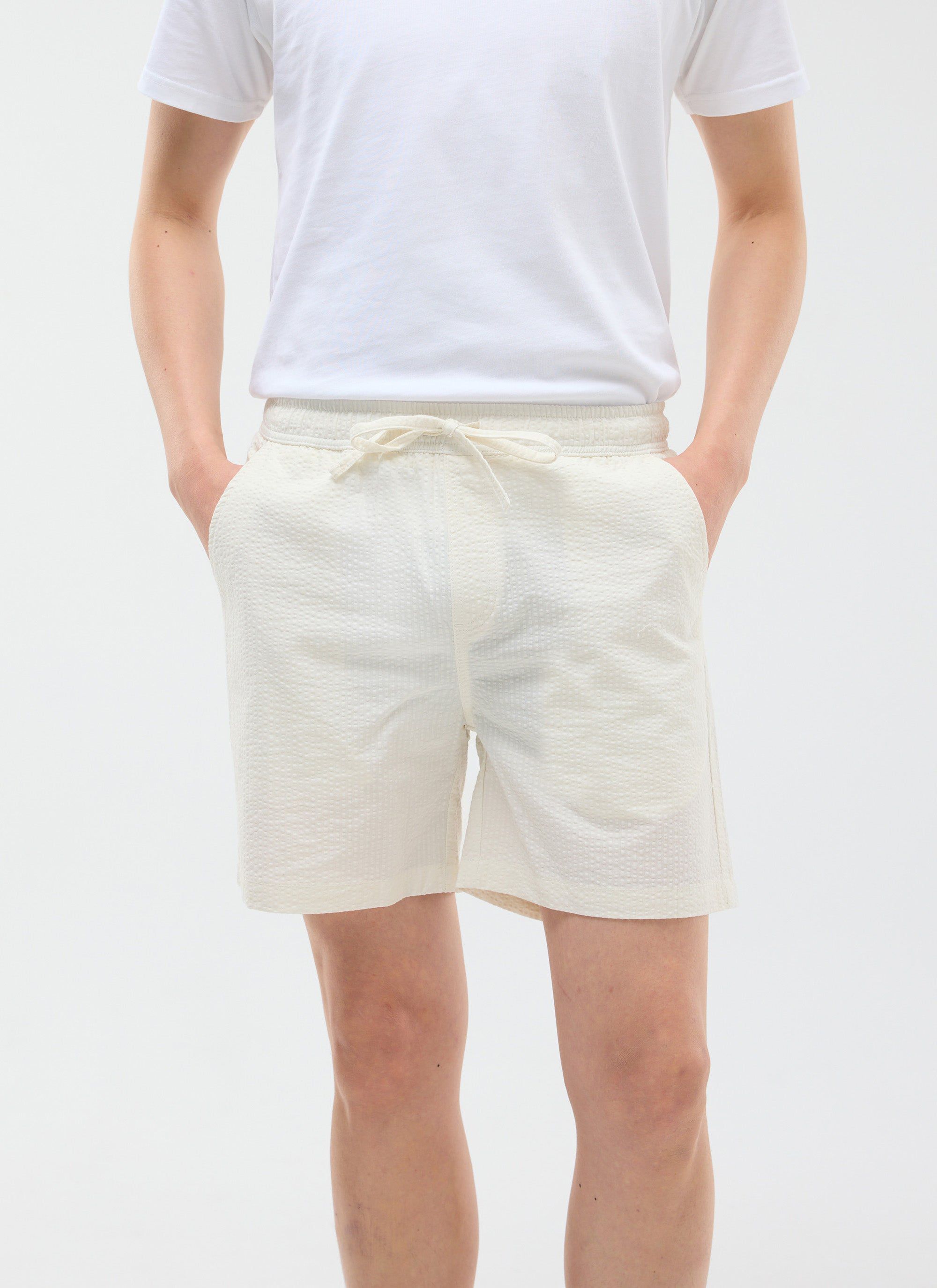 The Percival Classic Shorts in cream seersucker, shown on a model with a white t-shirt and hands in pockets.