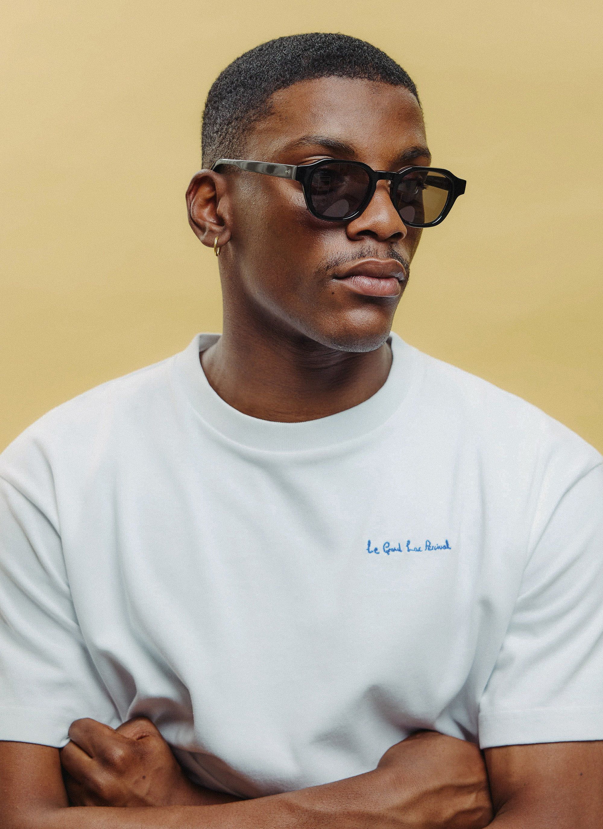 A model wears the Percival P001-E2 black sunglasses featuring polarised lenses, styled with a white tee on a yellow backdrop.
