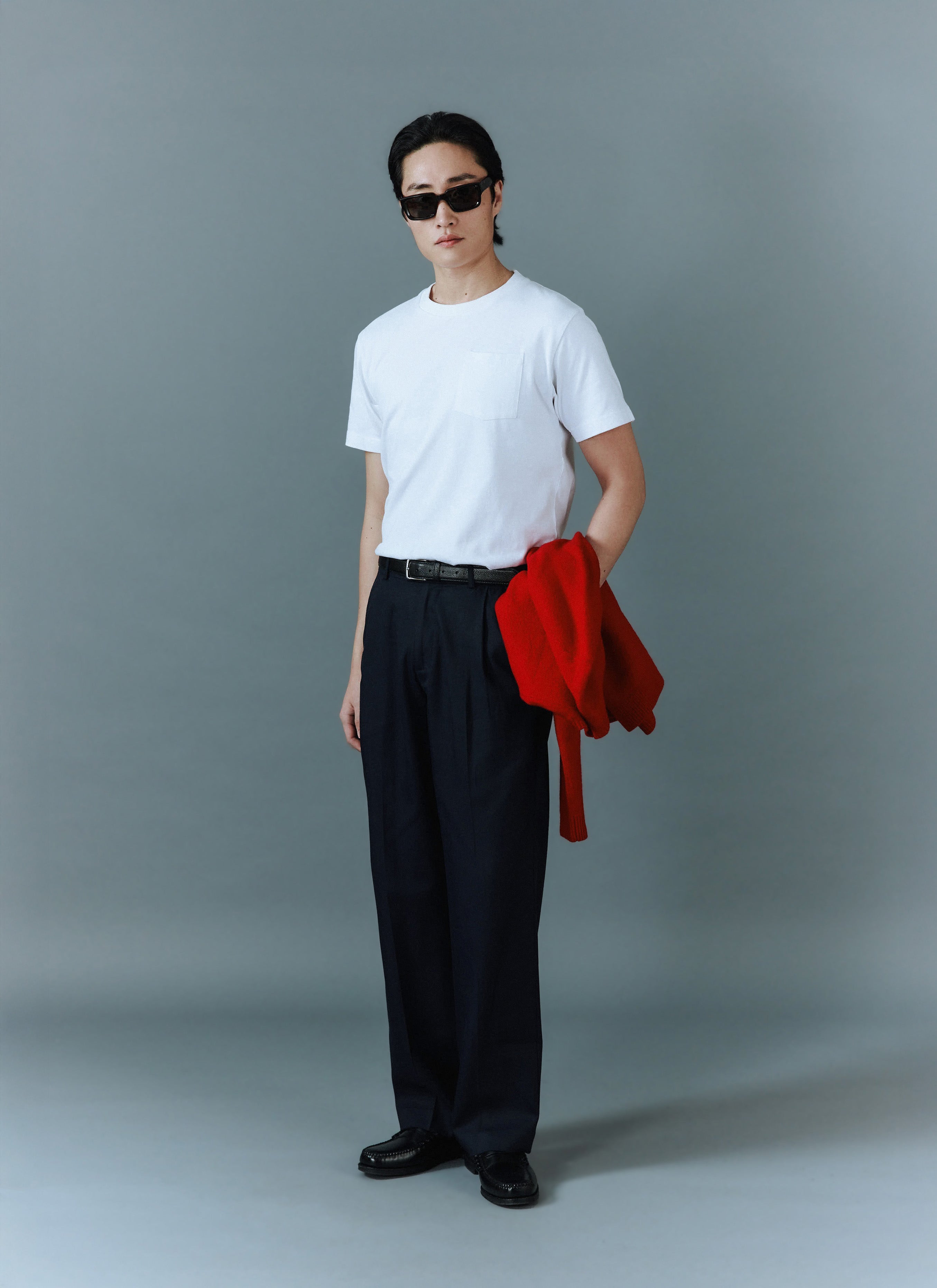 A man models the Percival white organic cotton Pocket T-Shirt, styled with dark trousers and holding a red jumper.