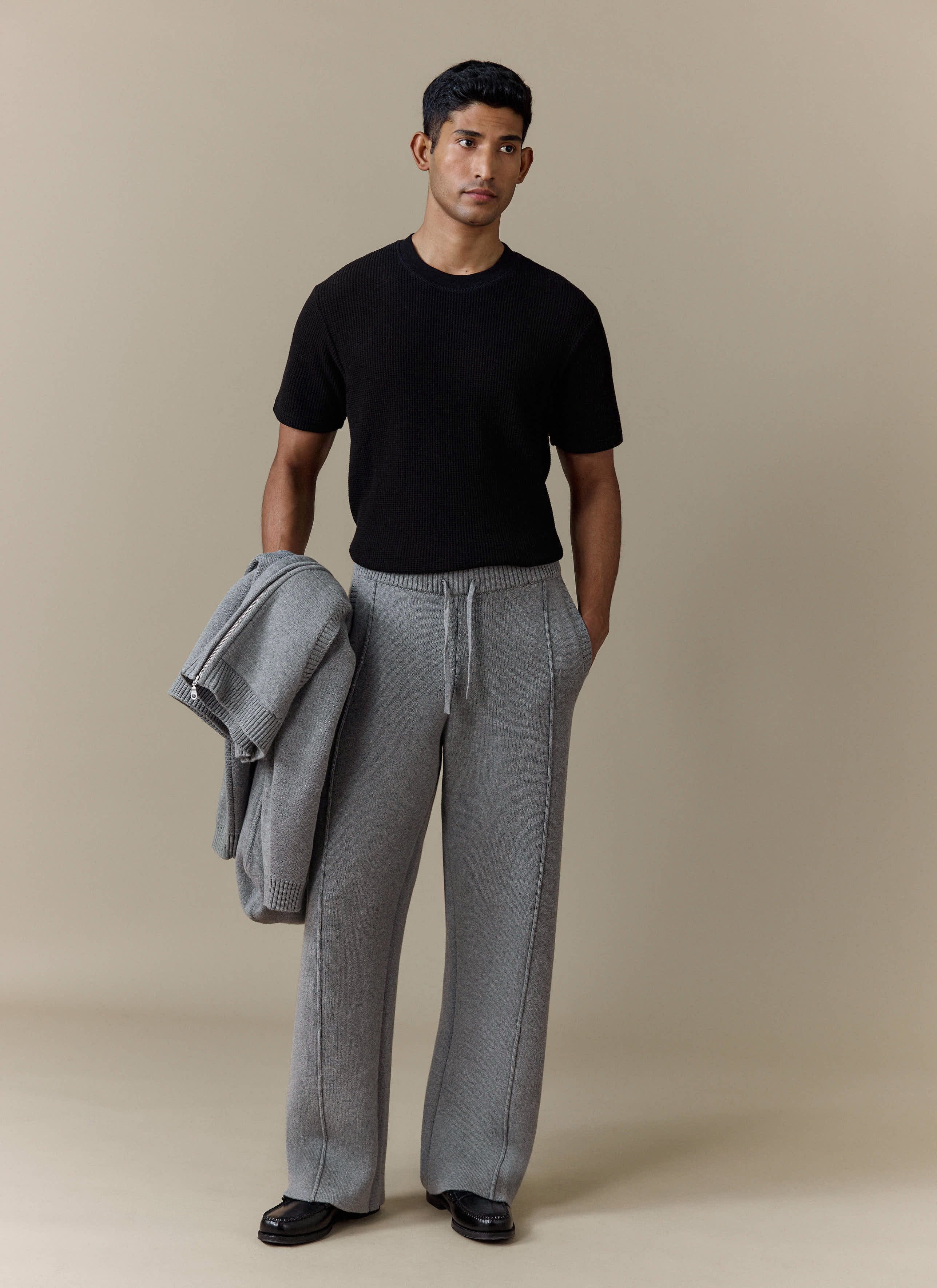A model wears the Percival Chronicle Knitted Straight Leg Trackpants in grey with a black tee, holding the matching hoodie.