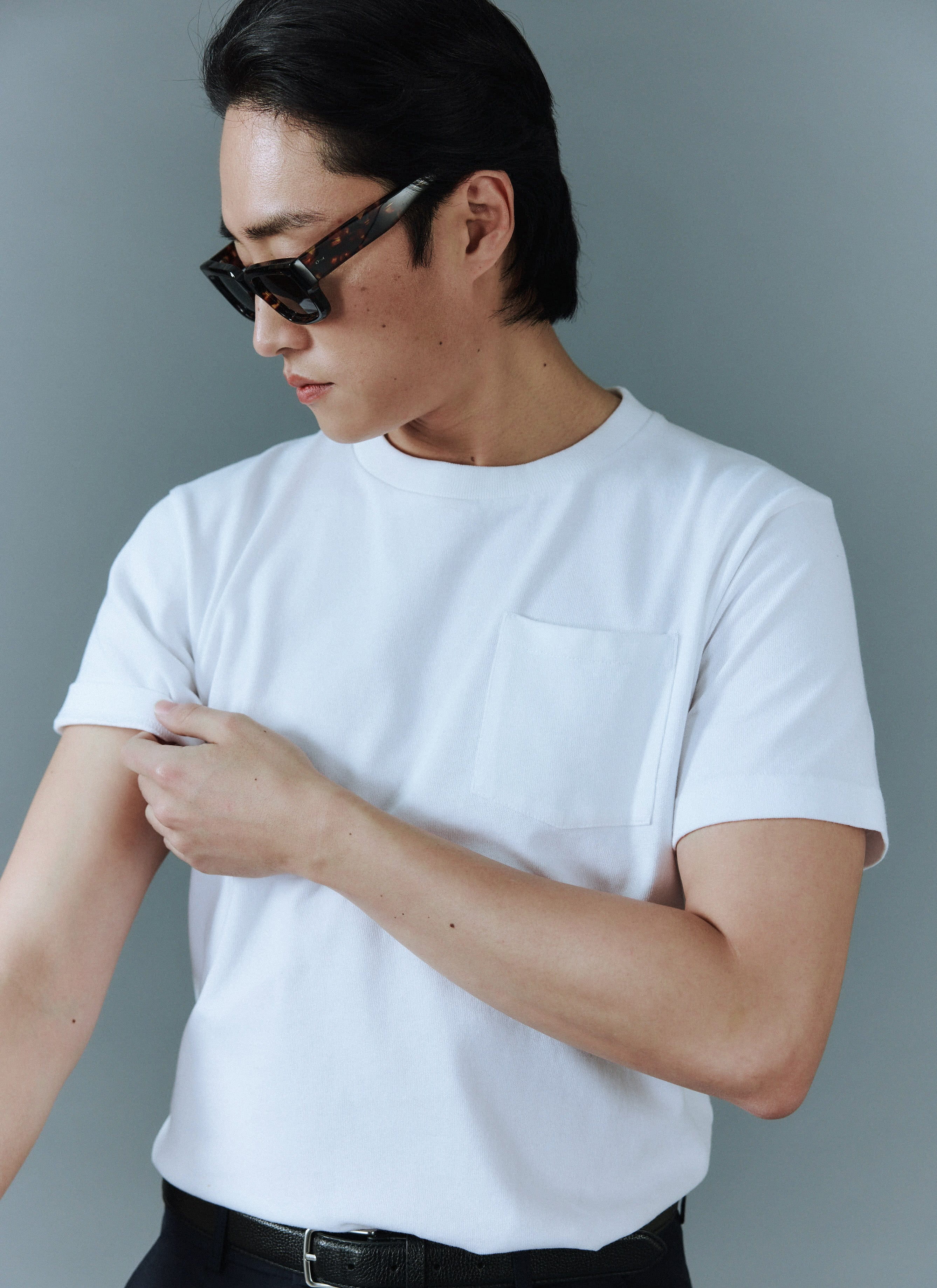 A man wearing sunglasses rolls up the sleeve of his white organic cotton Pocket T-Shirt from Percival.