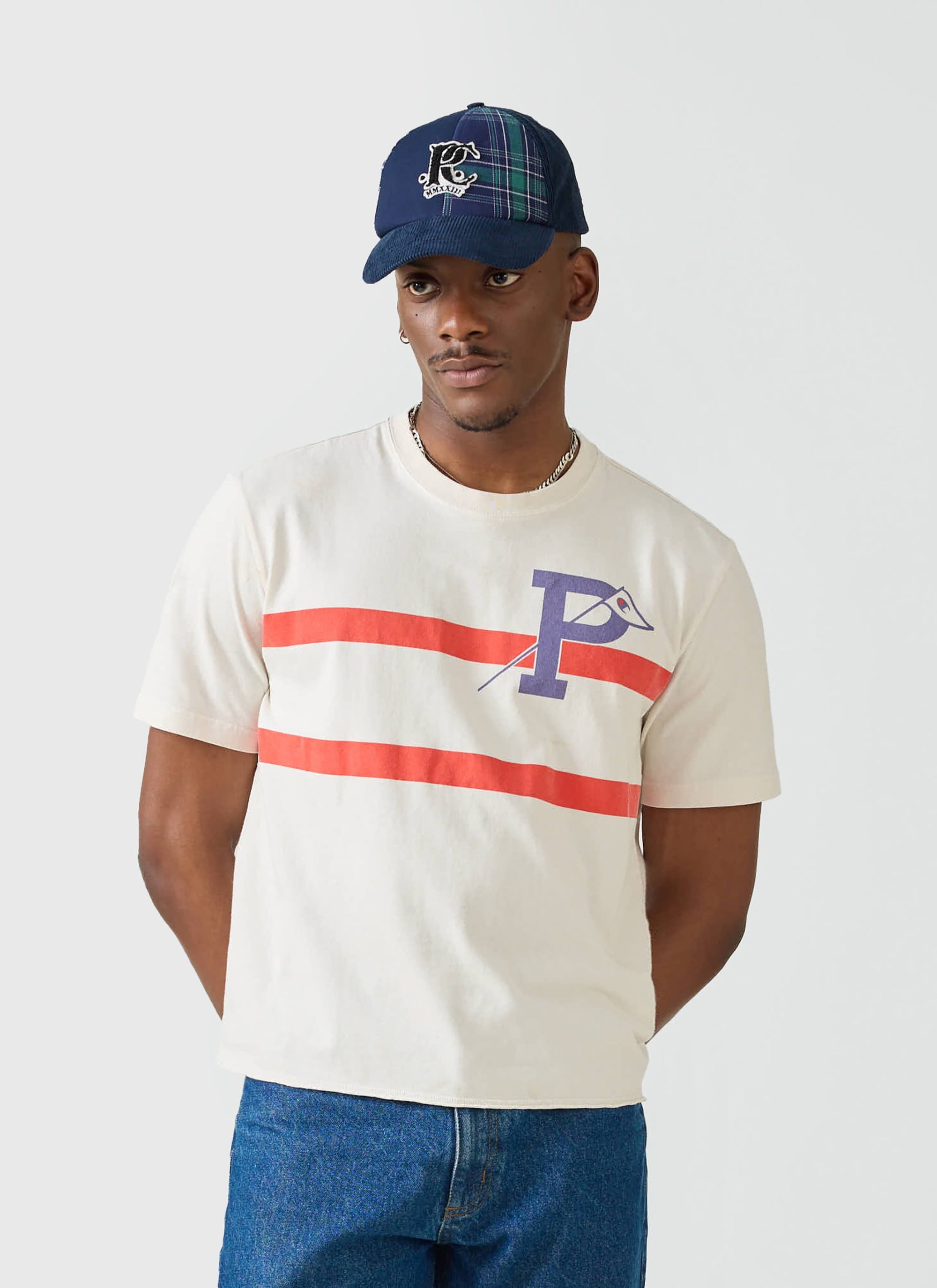 A model wears the ecru Champion x Percival Flag Stripe Raw Hem T Shirt, styled with blue jeans and a tartan cap.