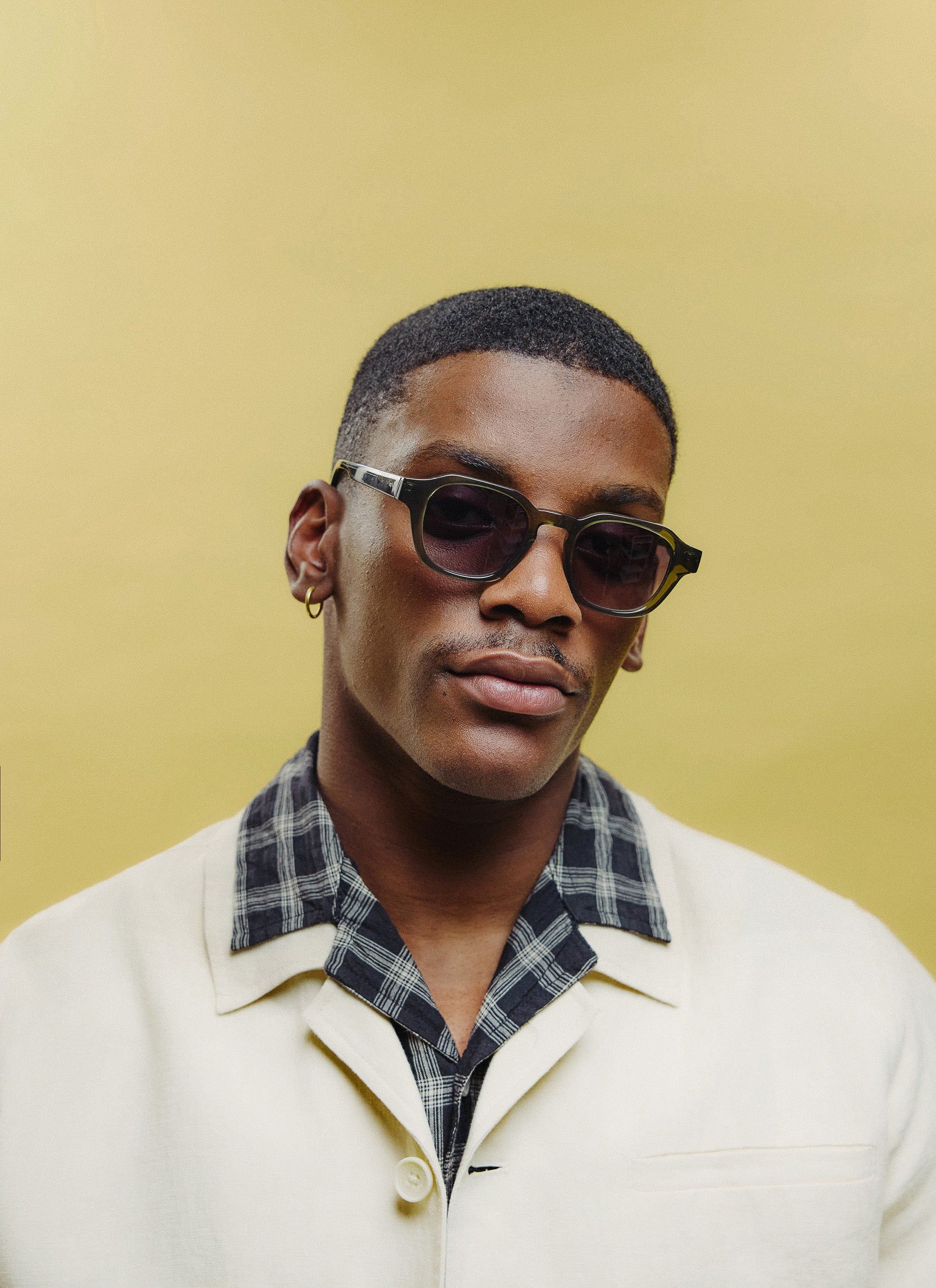 A model wears the Percival olive green sunglasses with a cream jacket and checked shirt against a yellow background.