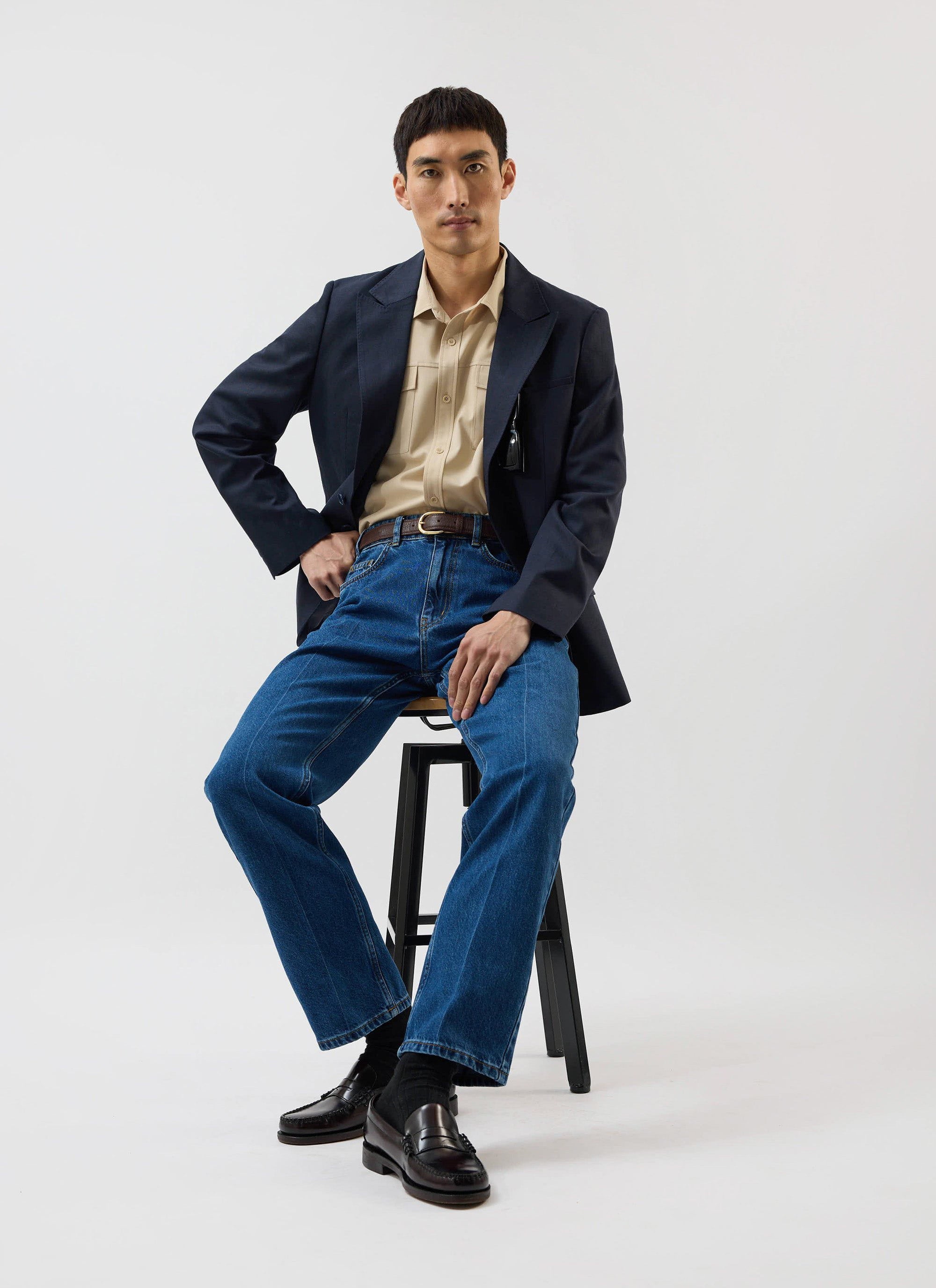 A model sits on a stool wearing the Percival x Warren Safari Shirt in camel beige, styled with a navy blazer and blue jeans.