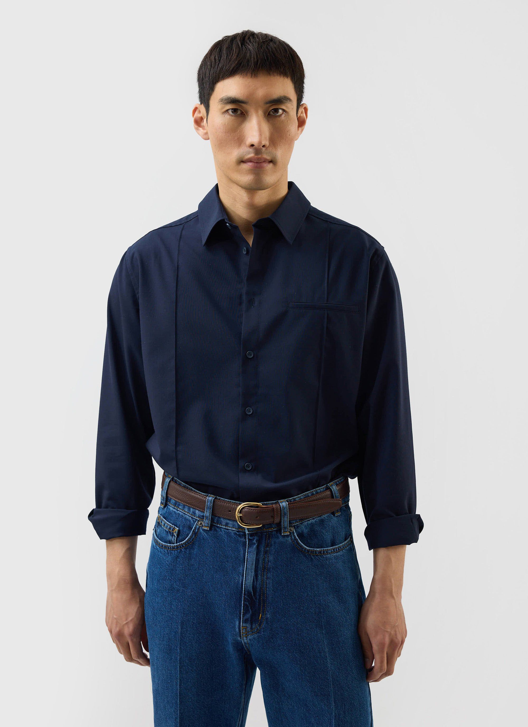 A model wears the Percival x Warren Pleated Shirt in navy blue, paired with classic blue jeans in a studio shot.
