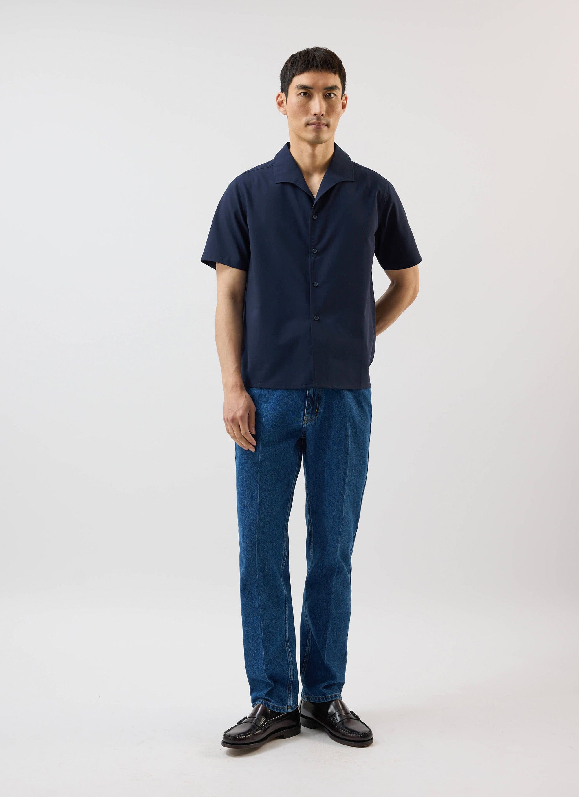 A man models the navy blue Percival x Warren Relaxed Cuban Drape Shirt, styled with dark wash jeans and black loafers.