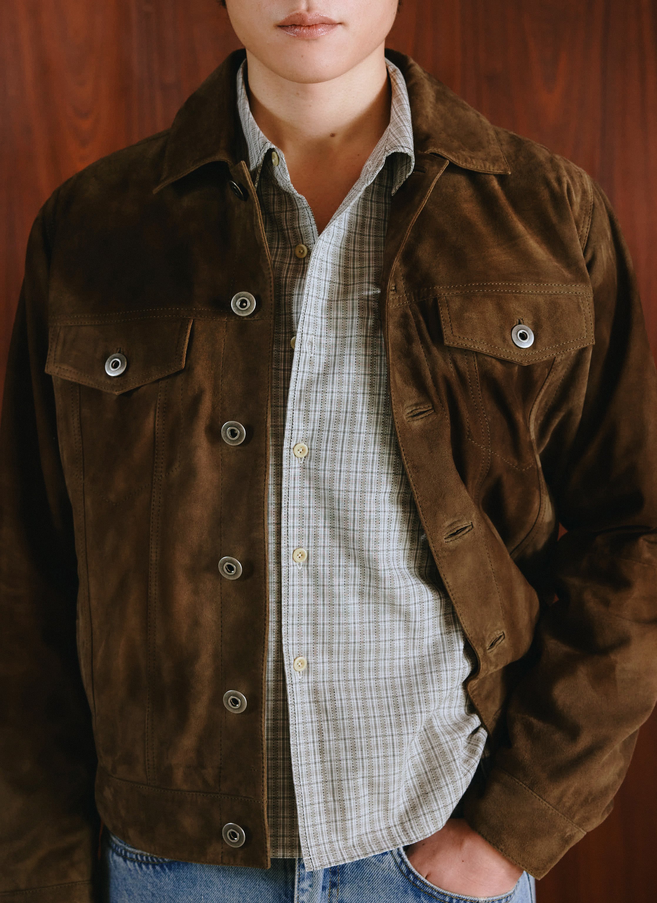 A model wears a brown suede Percival jacket over a checked shirt and blue jeans in a close-up studio shot.