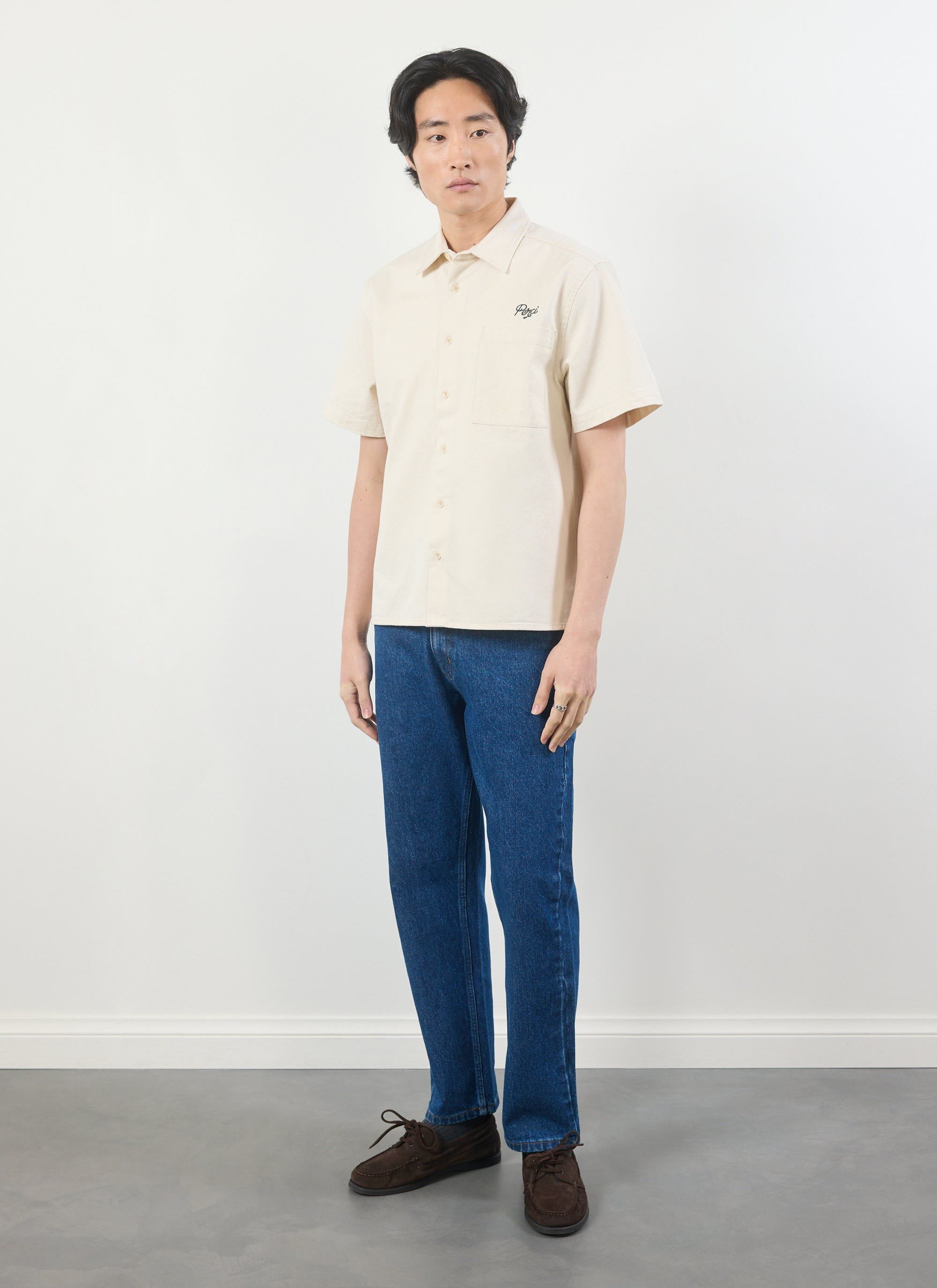 A model wears the Percival Olive Embroidered Cropped Shirt in white, styled with dark blue jeans and brown shoes.
