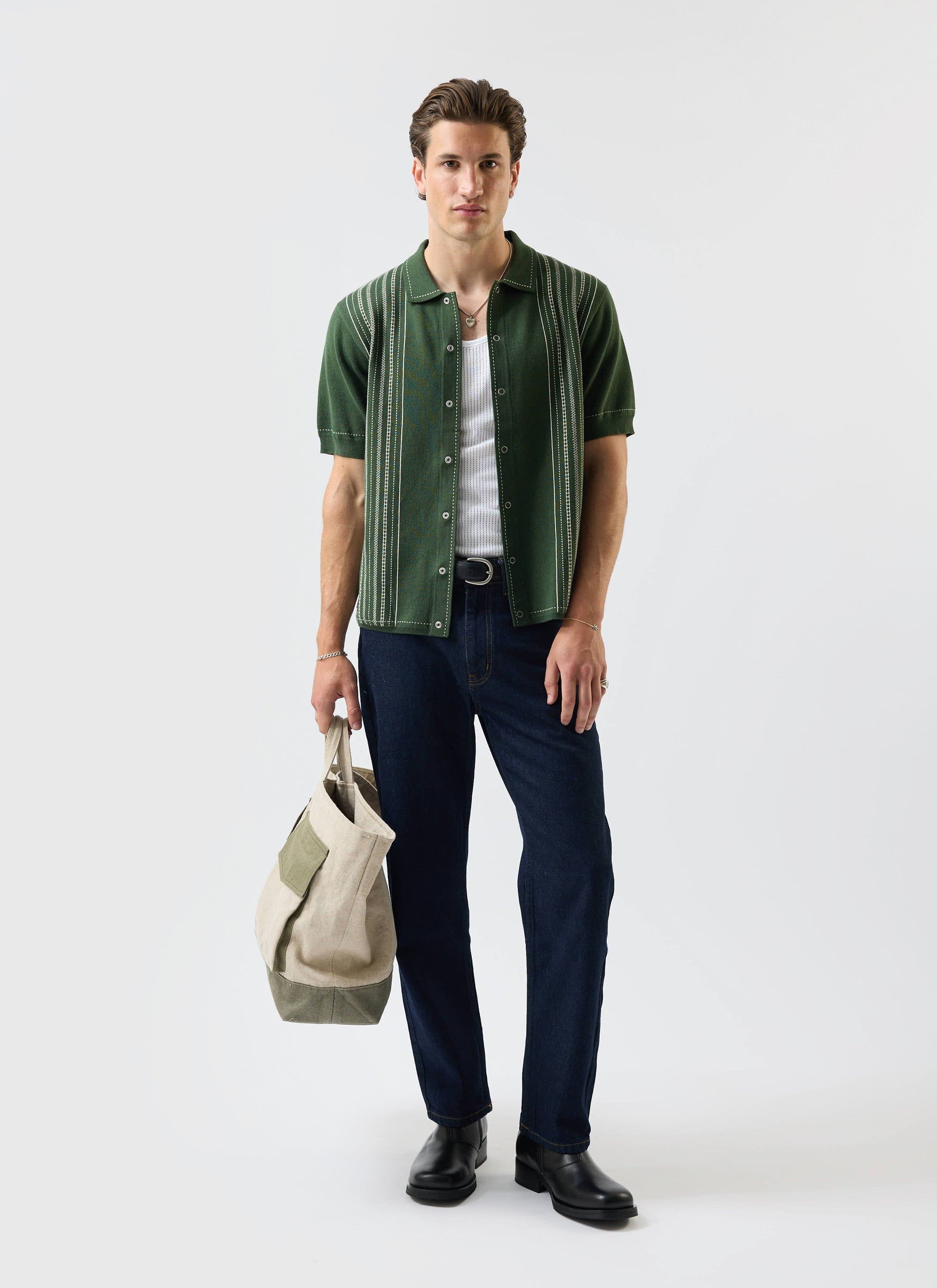 A model wears the Percival Accolades Knitted Shirt in forest green, open over a white vest with dark jeans and a tote bag.