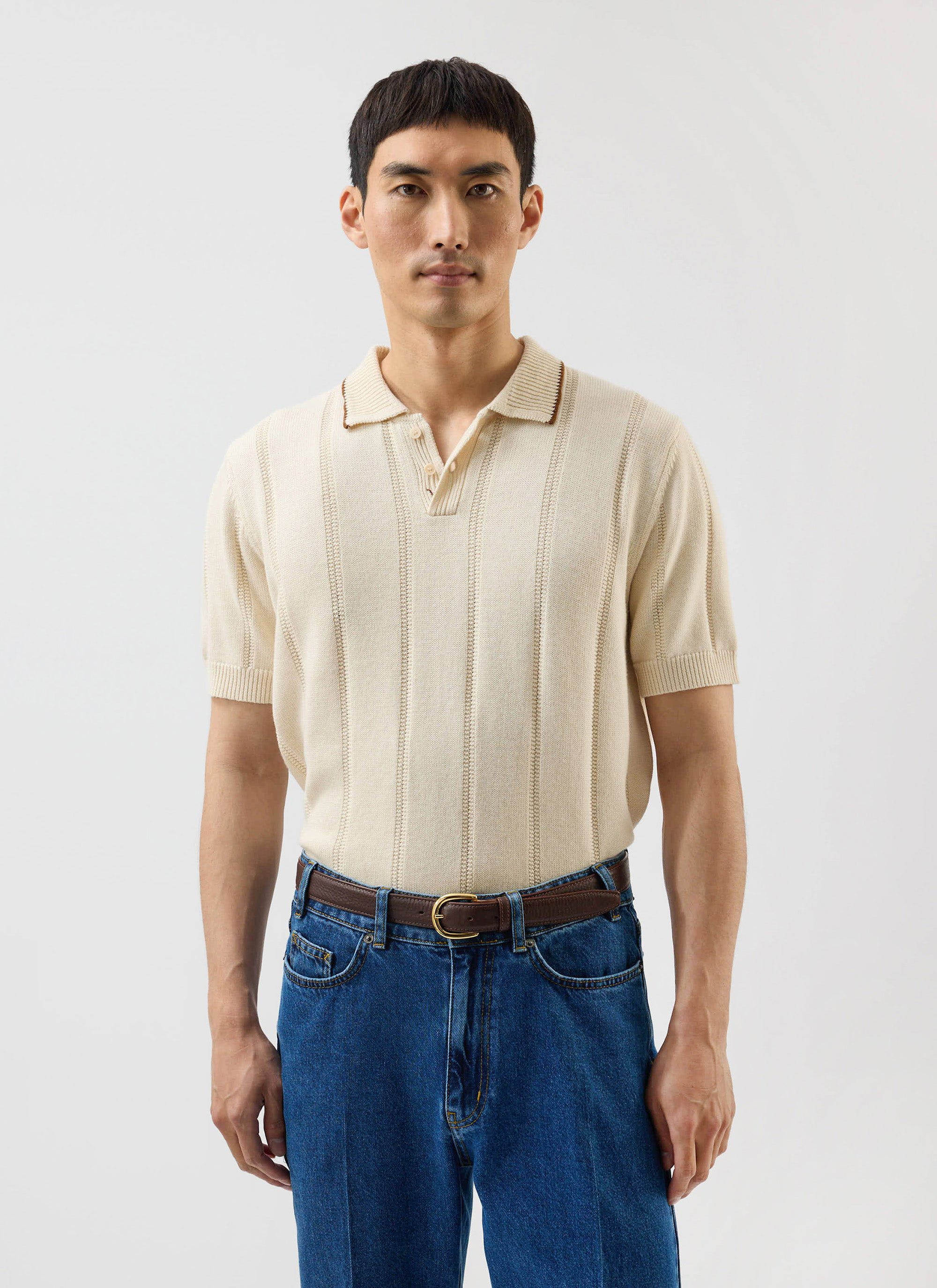 A man models the Percival x Warren ecru wool knitted polo with vertical stripes, tucked into blue jeans.