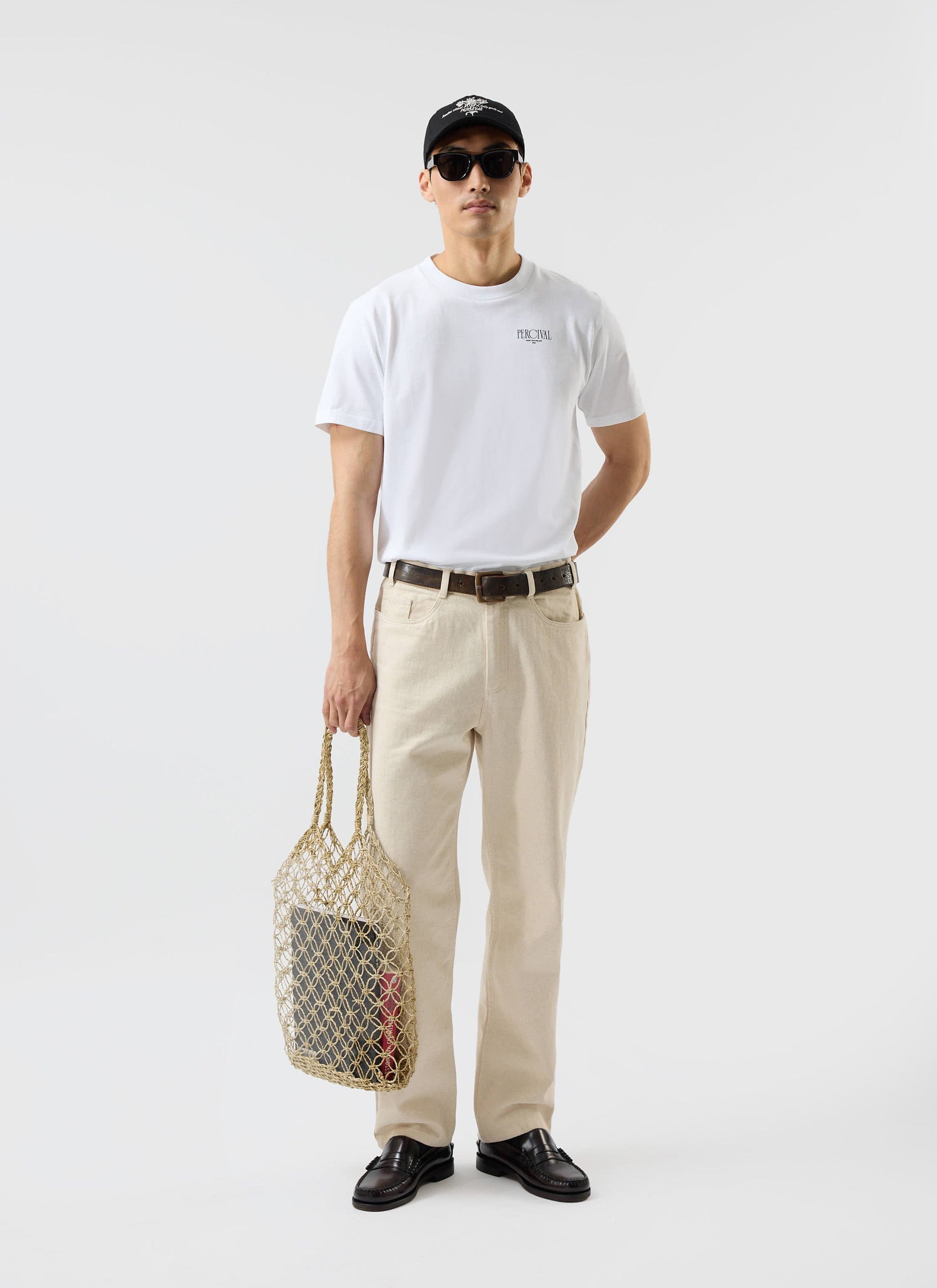 A model wears the white Percival Bistro T Shirt, styled with cream trousers, sunglasses, a cap, and holding a string bag.