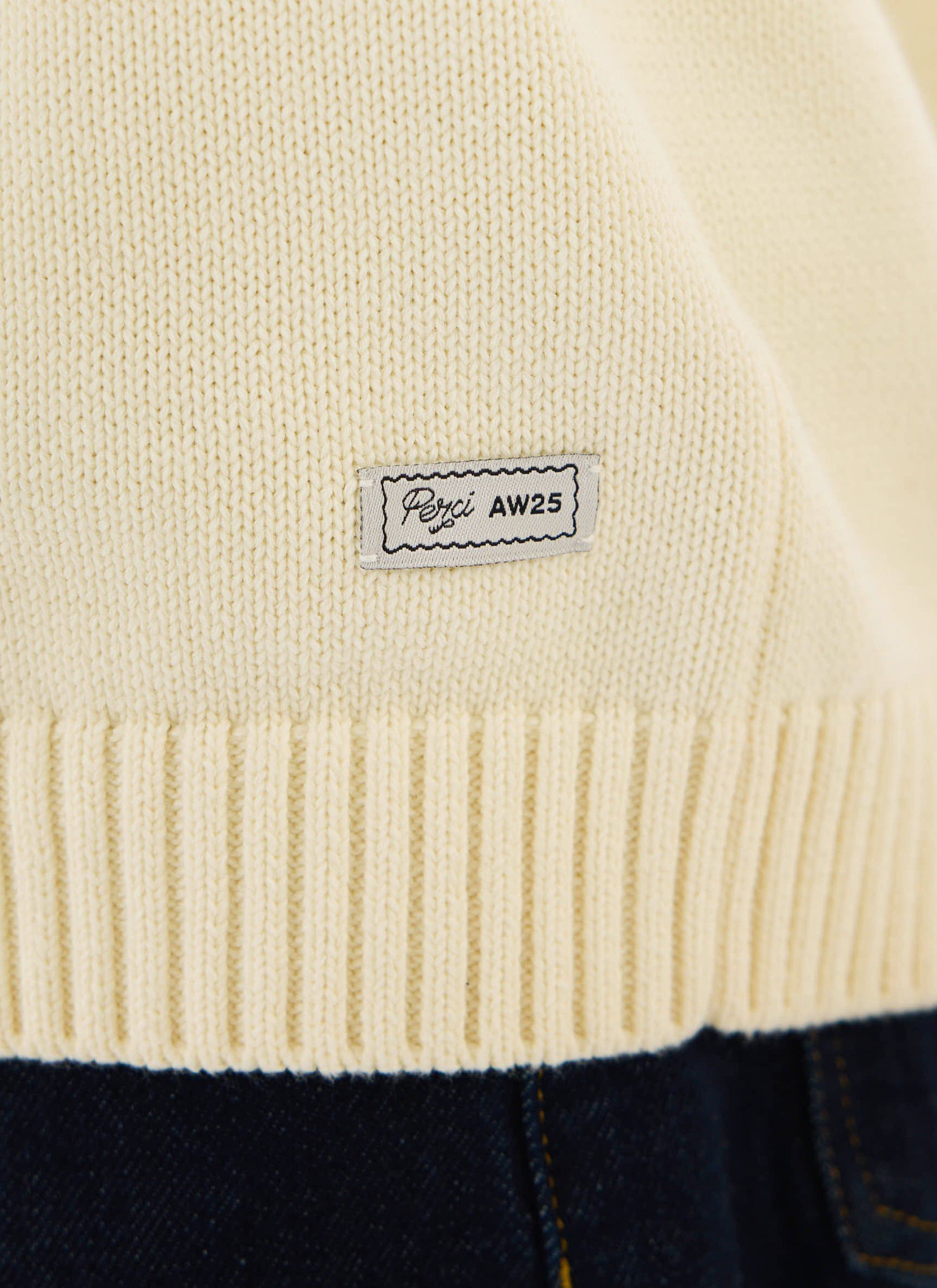 A close-up of the branded Percival AW25 patch on the ribbed hem of the ecru Verso Short Sleeve Knitted Shirt.