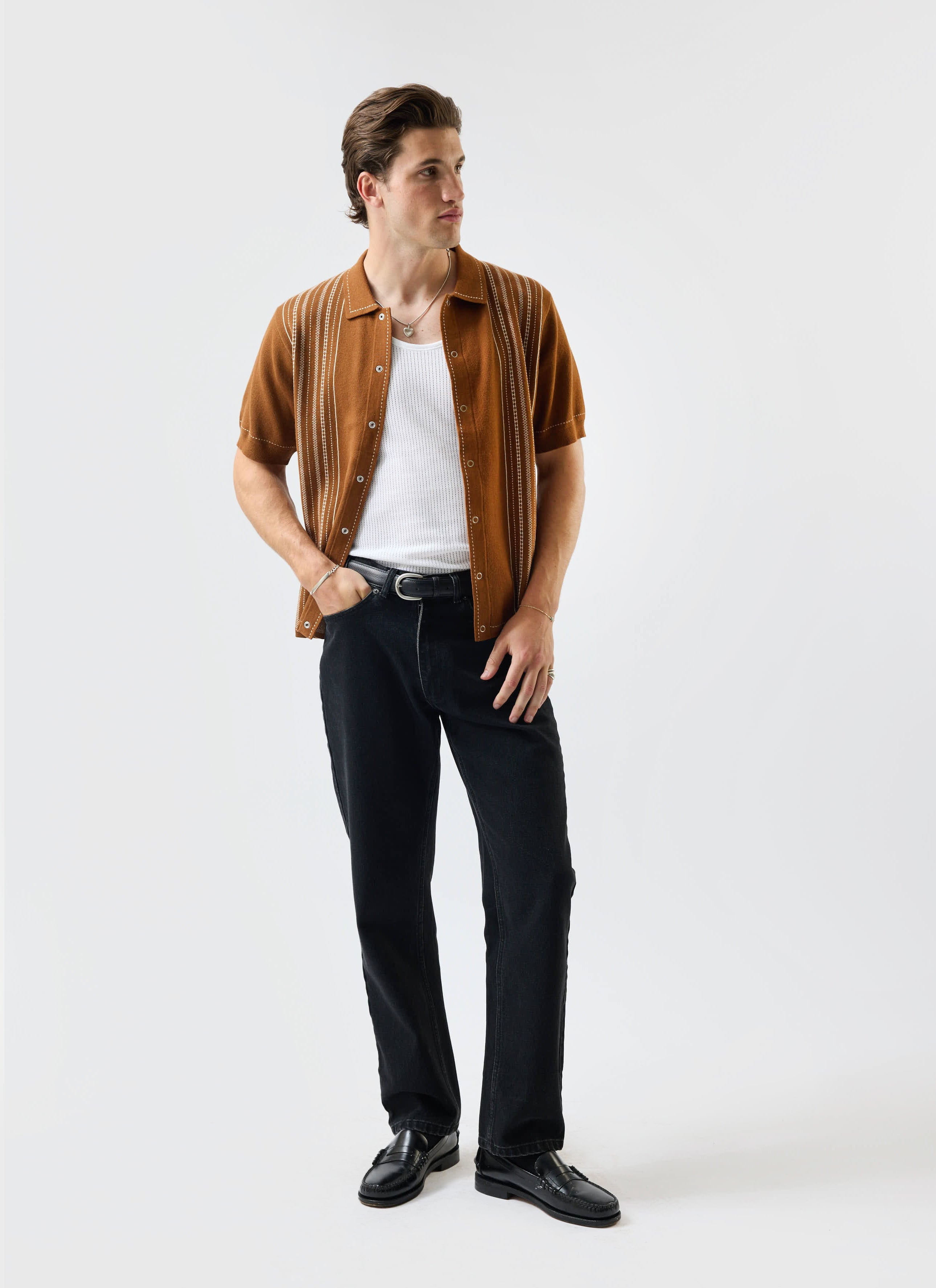 A man models the Percival Accolades Knitted Short Sleeve Shirt in tobacco brown, styled with black jeans and loafers.