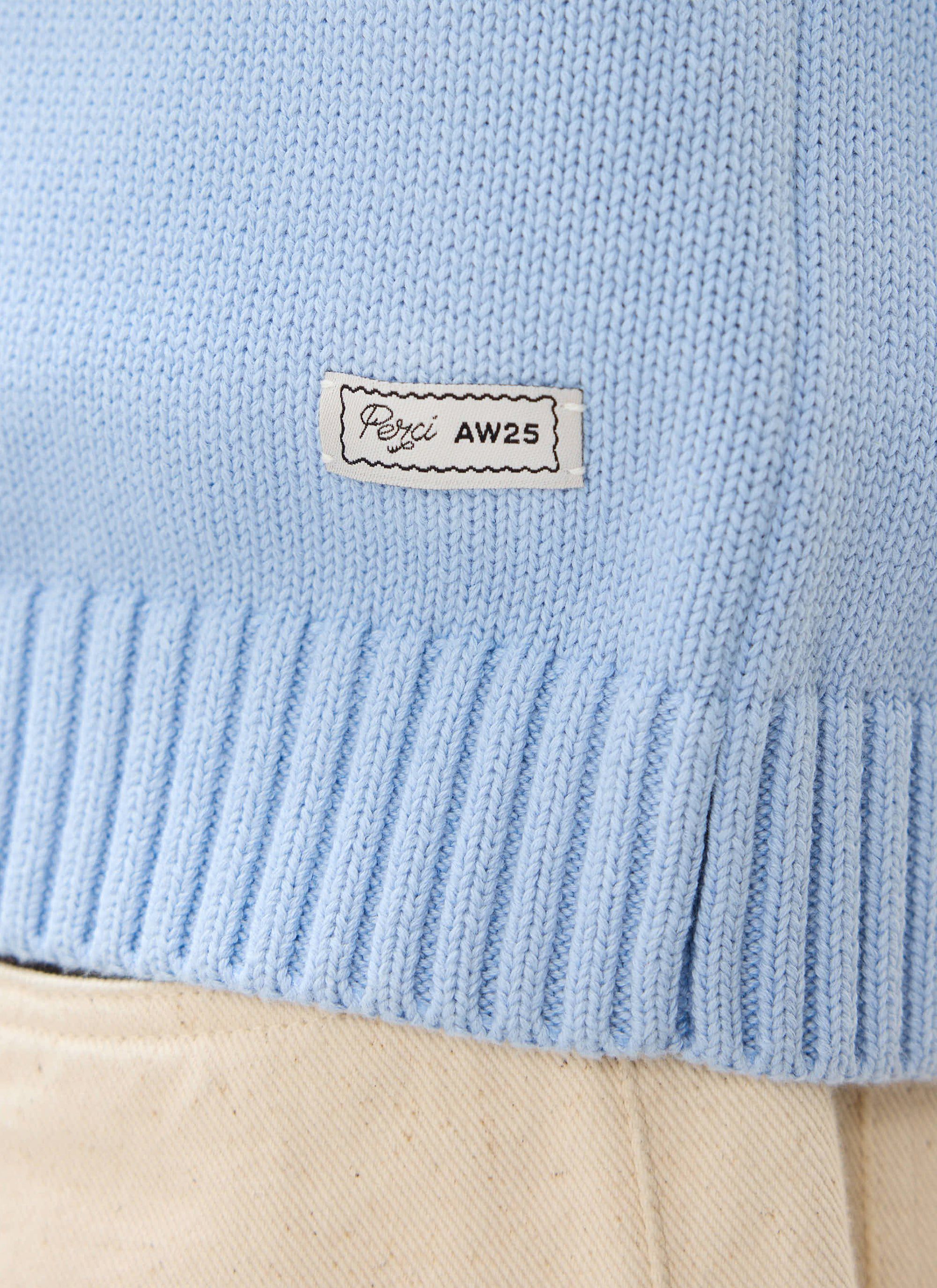 A close-up of the Percival Verso Short Sleeve Knitted Shirt in heather blue, showing the AW25 patch on the ribbed hem.