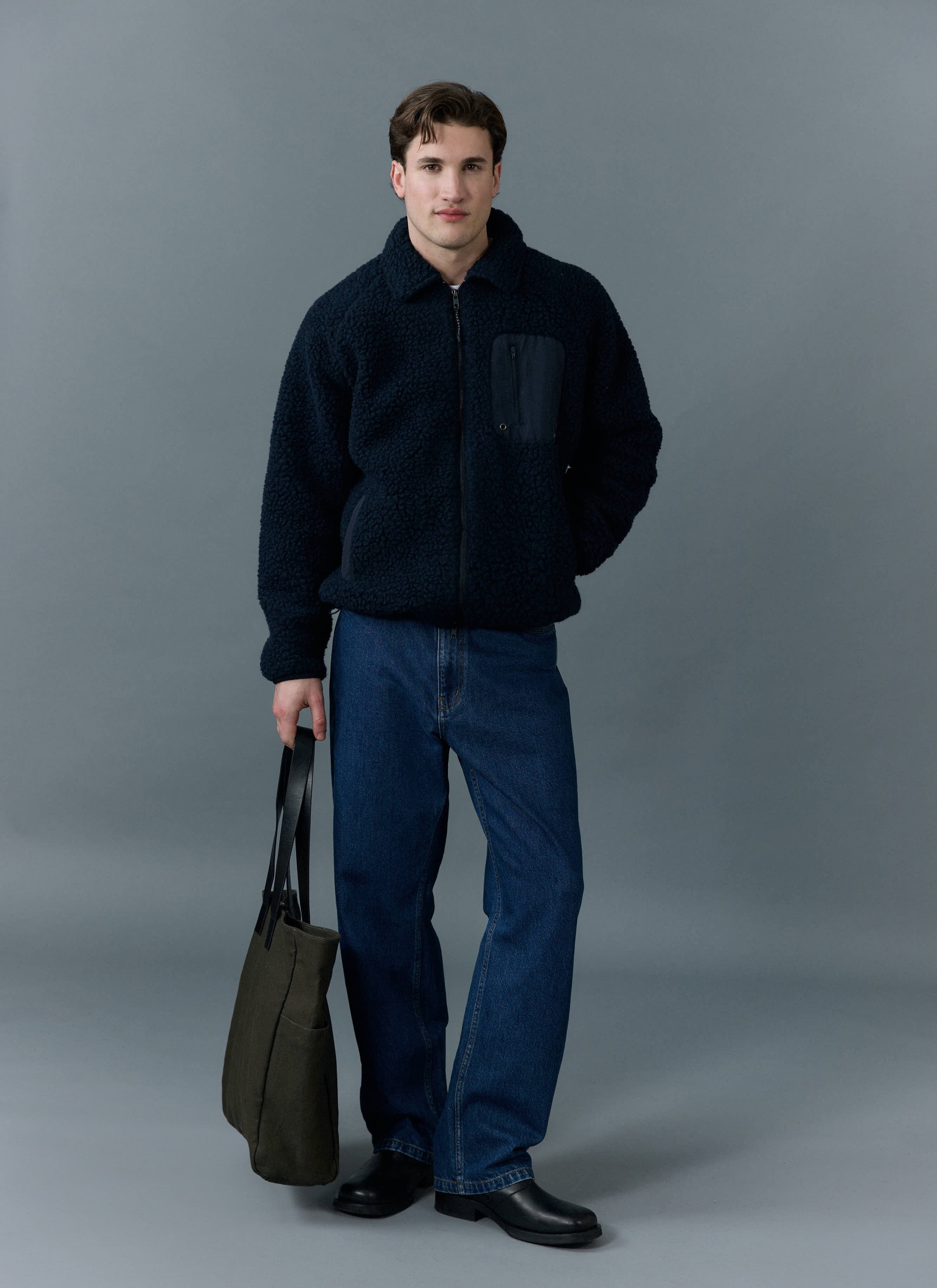 A man models the Percival Wide Leg Denim Jeans in mid wash blue, styled with a navy fleece jacket and a tote bag.