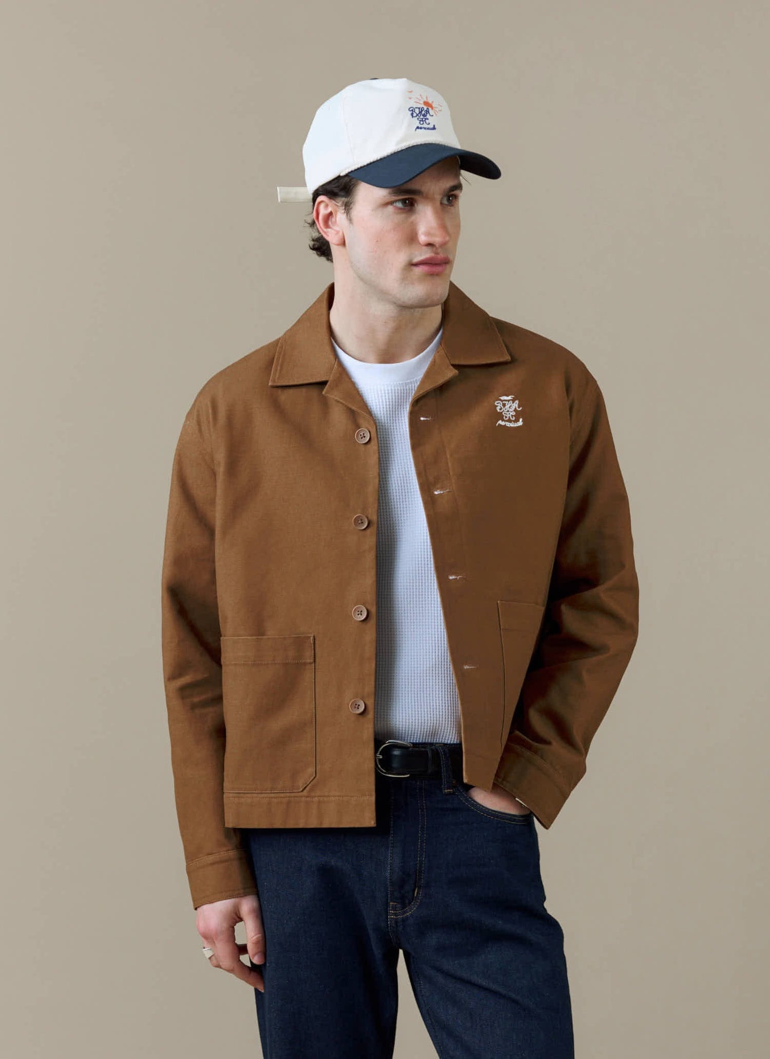 A studio shot of a model wearing the Percival x Brighton & Hove Albion F.C. brown Canvas Workshirt, a white t-shirt and cap.