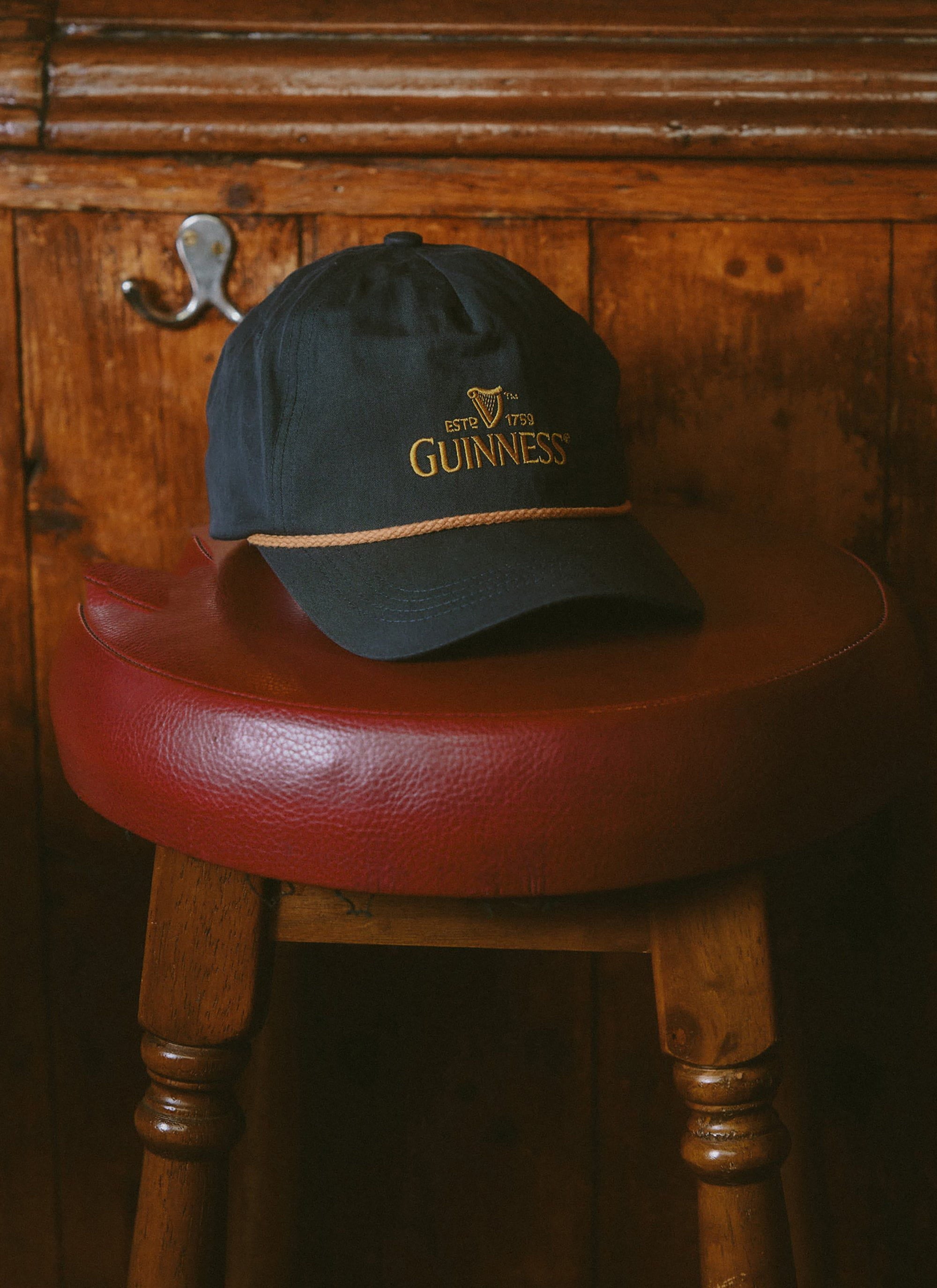 The navy Guinness x Percival cap from Percival resting on a red leather stool against a dark, wood-panelled wall.