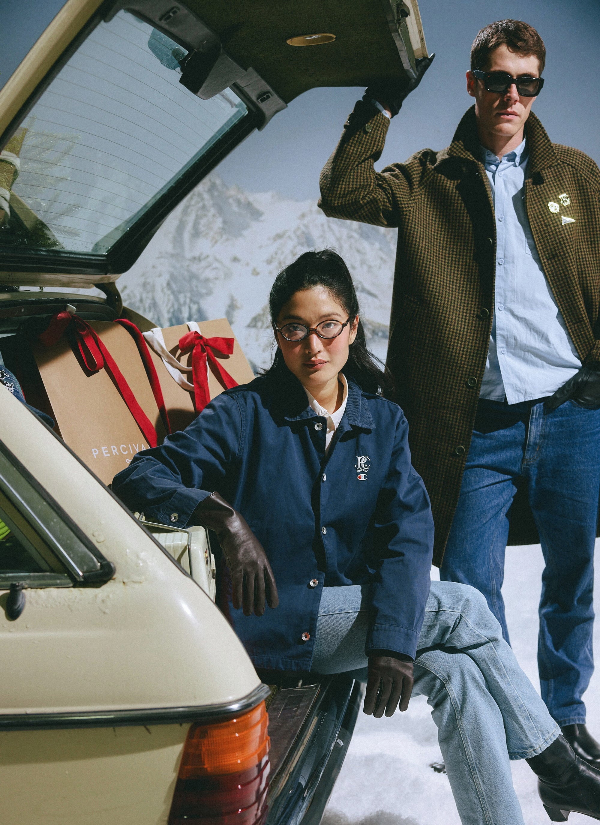 Models wear the Percival x Champion navy Cord Collar Jacket and blue Oxford Shirt in a snowy, après-ski scene with a car.