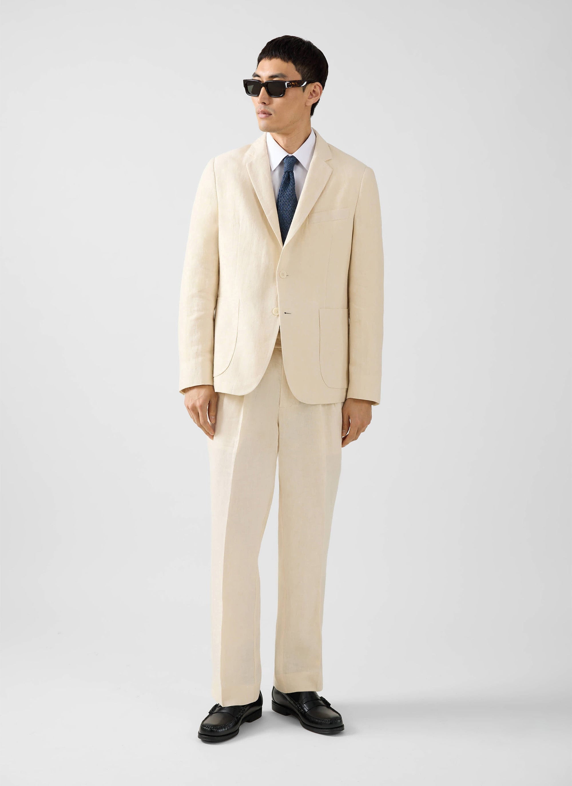 A model wears the full Percival tailored linen suit in cream, styled with a shirt, tie, and sunglasses.
