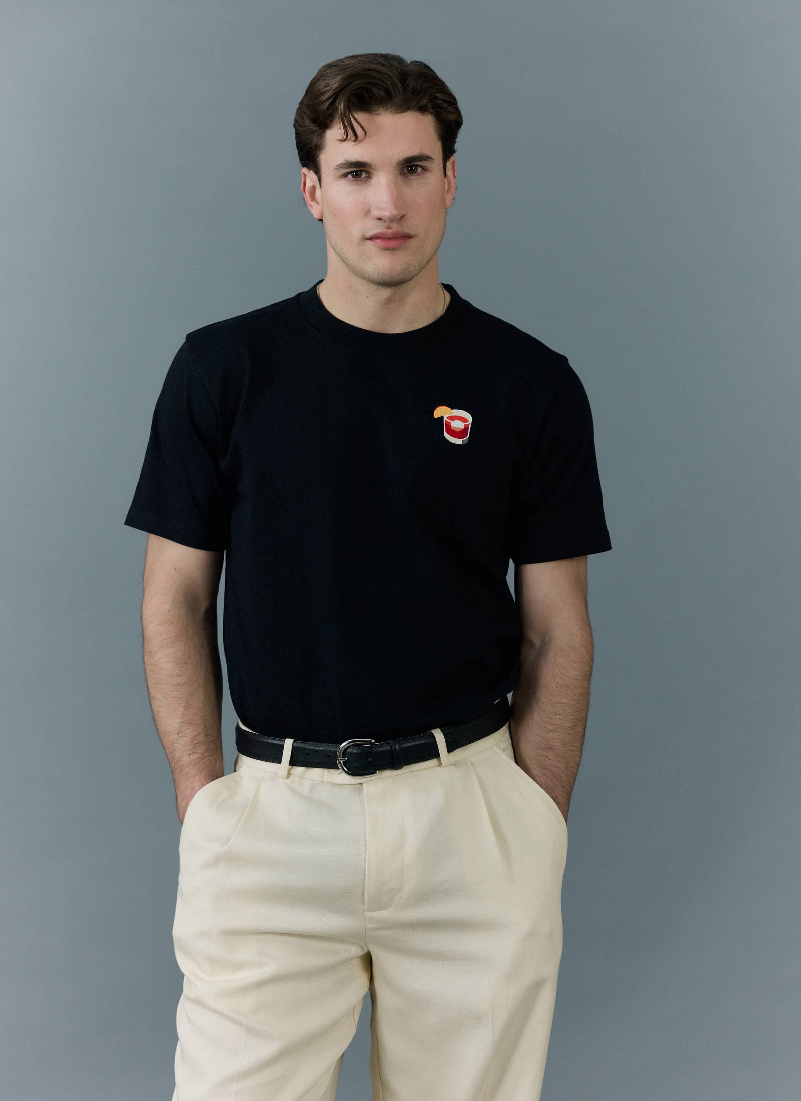 A model wears the black Percival Negroni T-shirt with its signature embroidered cocktail, styled with cream trousers.