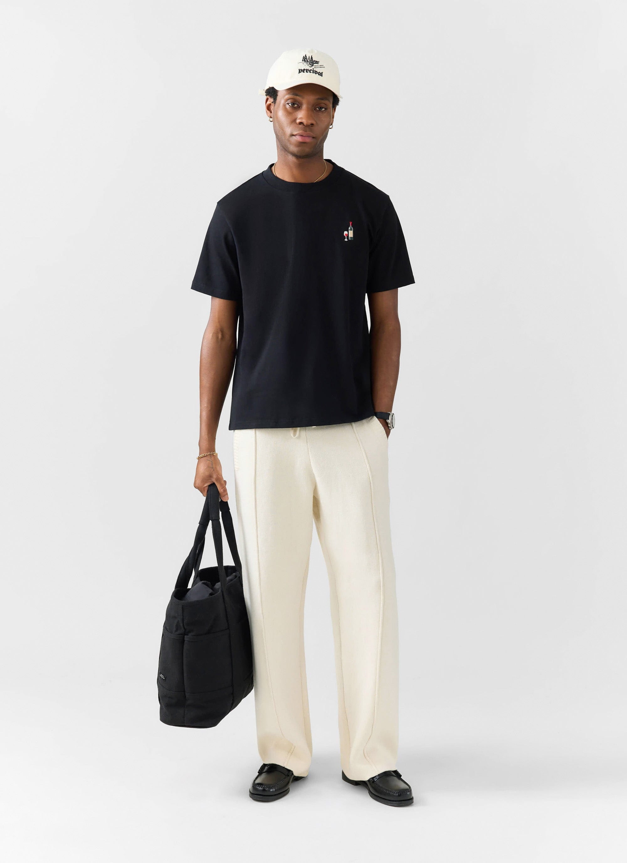 A model wears the Percival black Red Wine Embroidered T-shirt, styled with cream trousers, a white cap and a black tote bag.