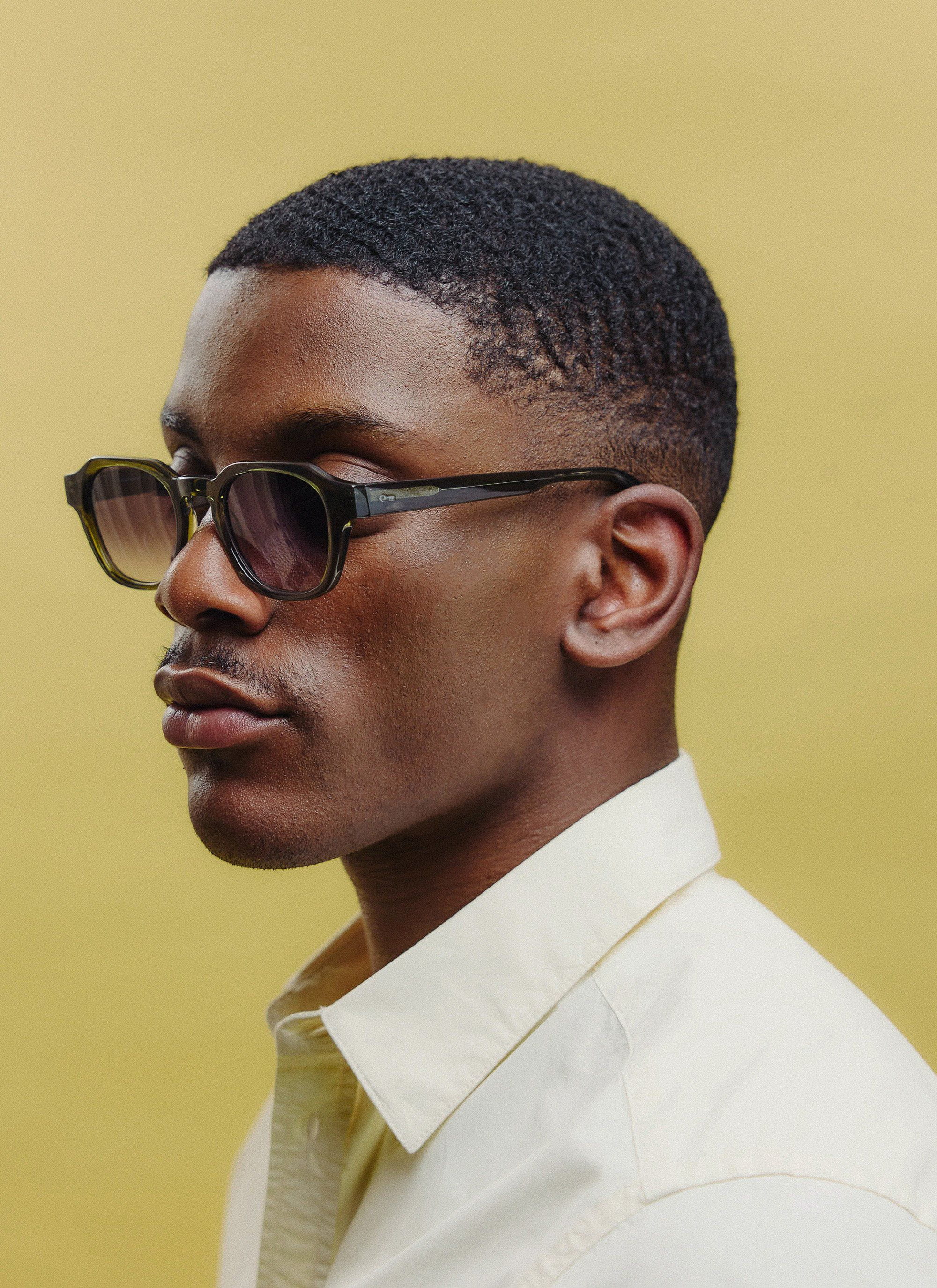 A close-up studio shot of a model wearing Percival's olive green sunglasses with light brown lenses, viewed from the side.