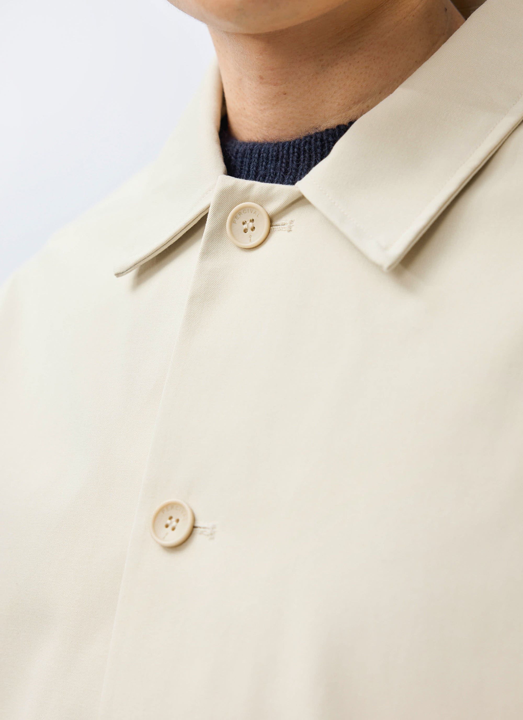 A close-up of the Percival Sherlock Short Trench Coat in stone, showing the collar and branded buttons over a blue knit.