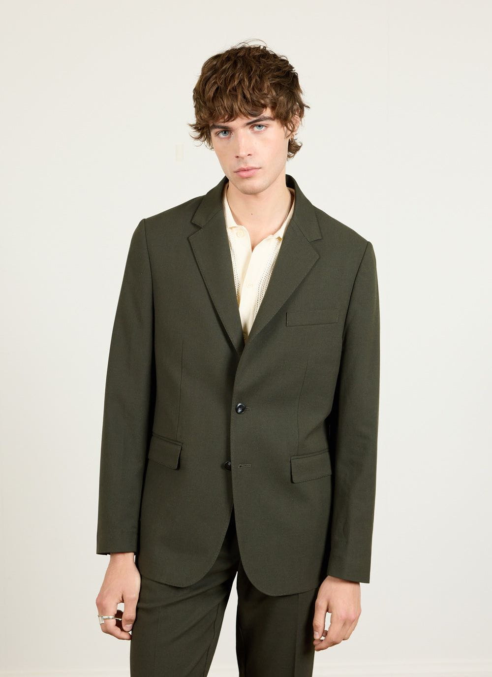 A model wears the Percival Tailored Blazer in forest green wool with a cream polo shirt against a plain studio background.